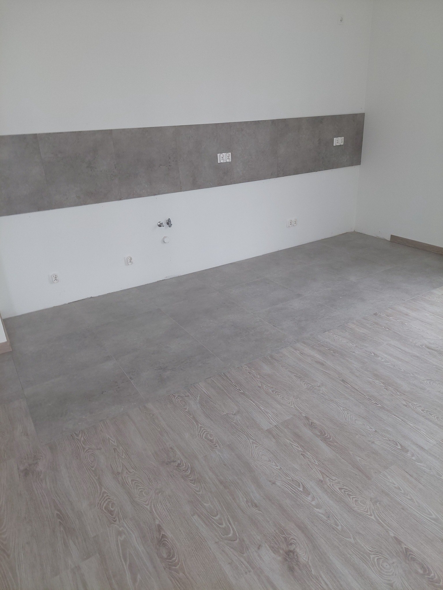 Wykończone wnętrze: szare płytki na ścianie i podłodze przechodzące w panele podłogowe o jasnym, drewnopodobnym wzorze. Widoczne gniazdka elektryczne i przyłącza wodne.