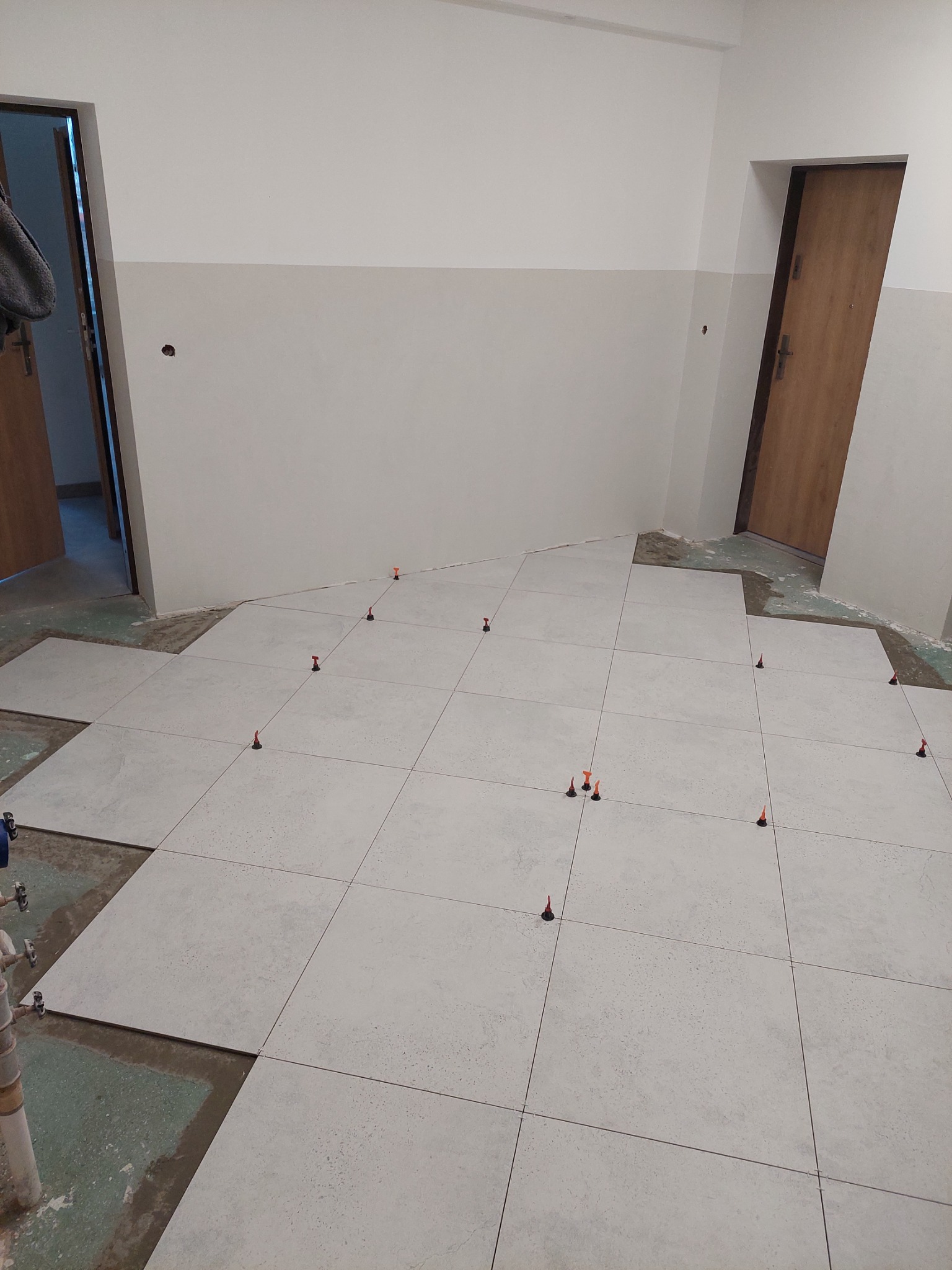 Układanie jasnych, kwadratowych płytek podłogowych z użyciem poziomujących klipsów i klinów w pomieszczeniu z dwukolorowymi ścianami i drewnianymi drzwiami.