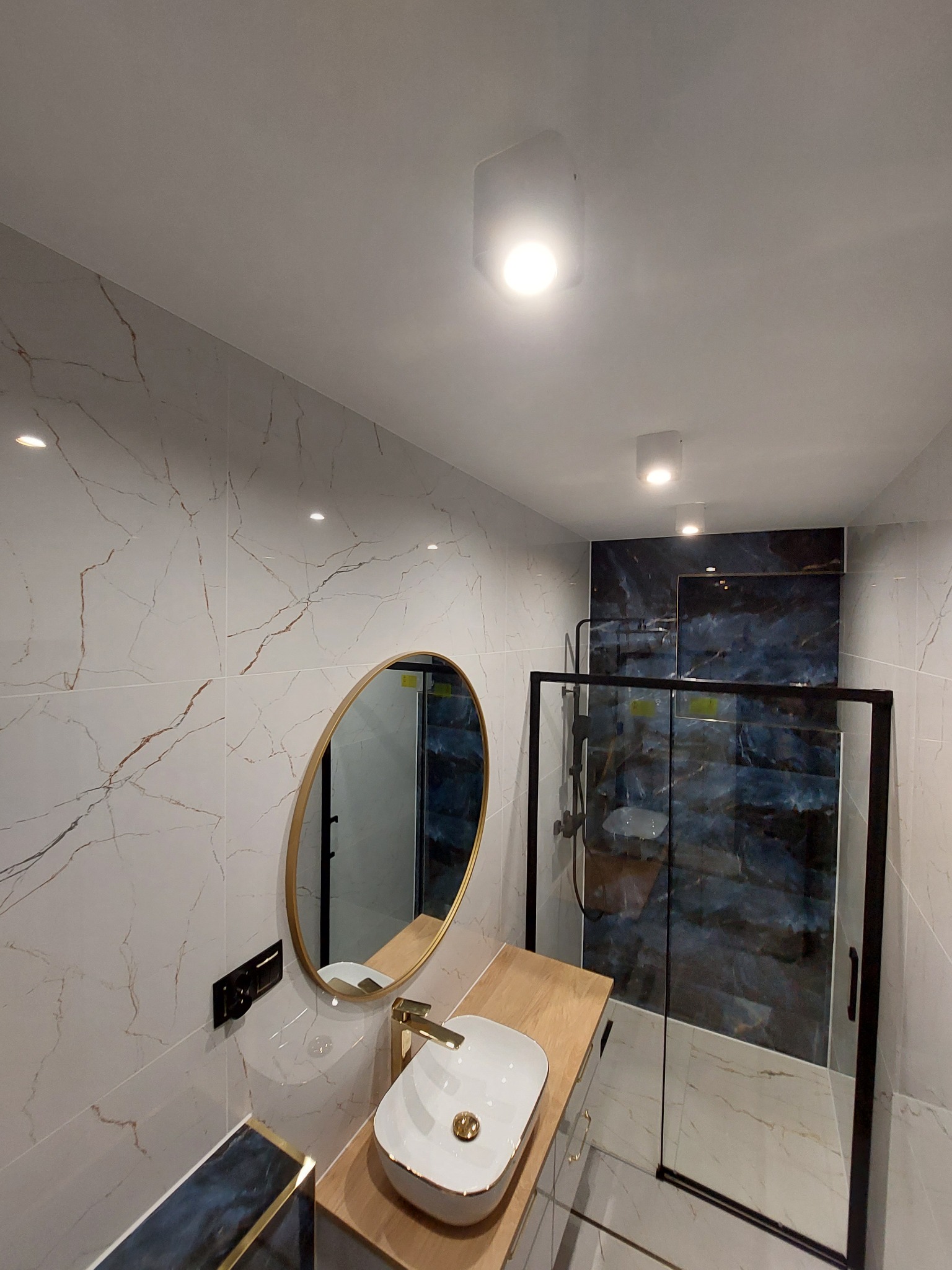 Nowoczesna łazienka z marmurowymi płytkami, owalnym lustrem w złotej ramie, umywalką nablatową i kabiną prysznicową z czarnymi profilami.