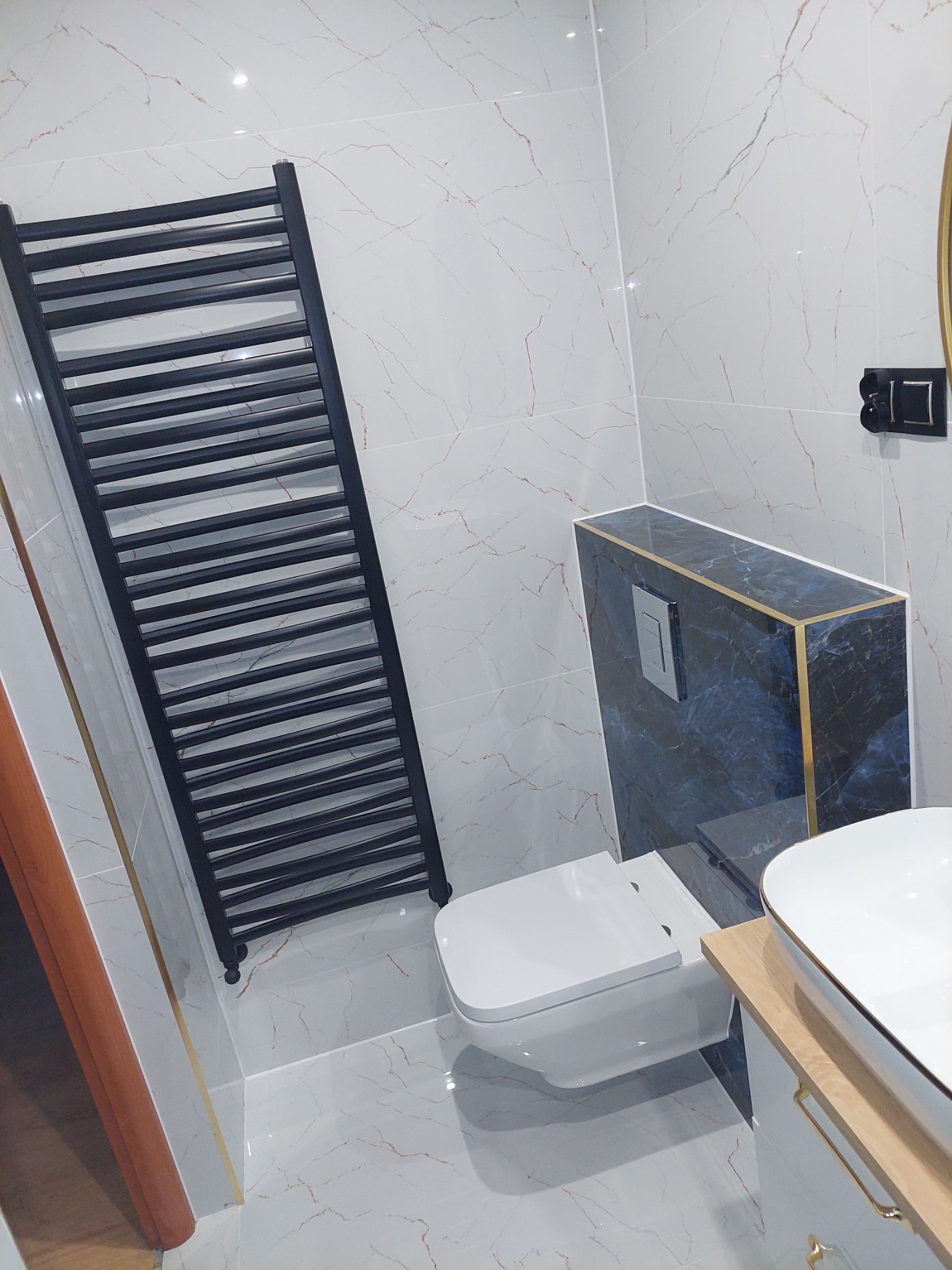 Nowoczesna łazienka z białymi marmurkowymi płytkami, czarnym grzejnikiem drabinkowym, podwieszaną toaletą i umywalką nablatową z elementami w kolorze złota.
