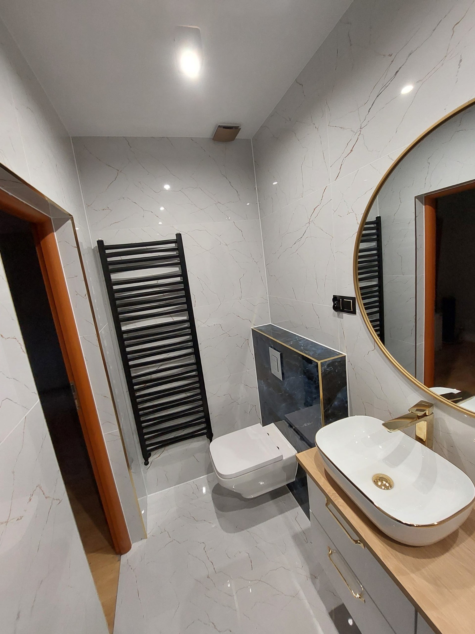 Nowoczesna łazienka z marmurowymi płytkami, podwieszaną toaletą w zabudowie z ciemnego marmuru ze złotymi akcentami, owalną umywalką nablatową z baterią w kolorze złota, czarnym grzejnikiem...
