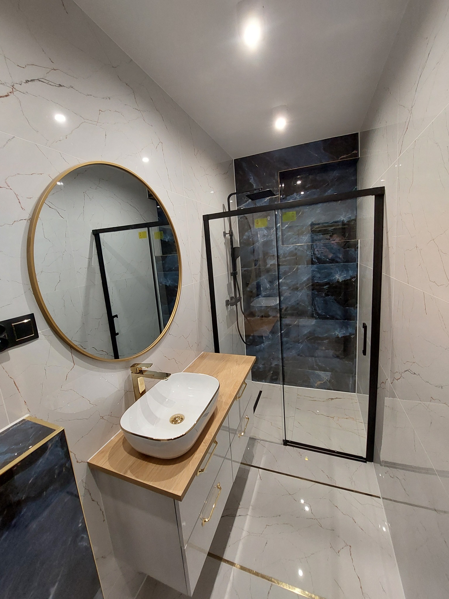Nowoczesna łazienka z kabiną prysznicową typu walk-in z czarnymi ramami, okrągłym lustrem w złotej ramie nad białą szafką z umywalką nablatową oraz marmurowymi płytkami na ścianach i podłodze.