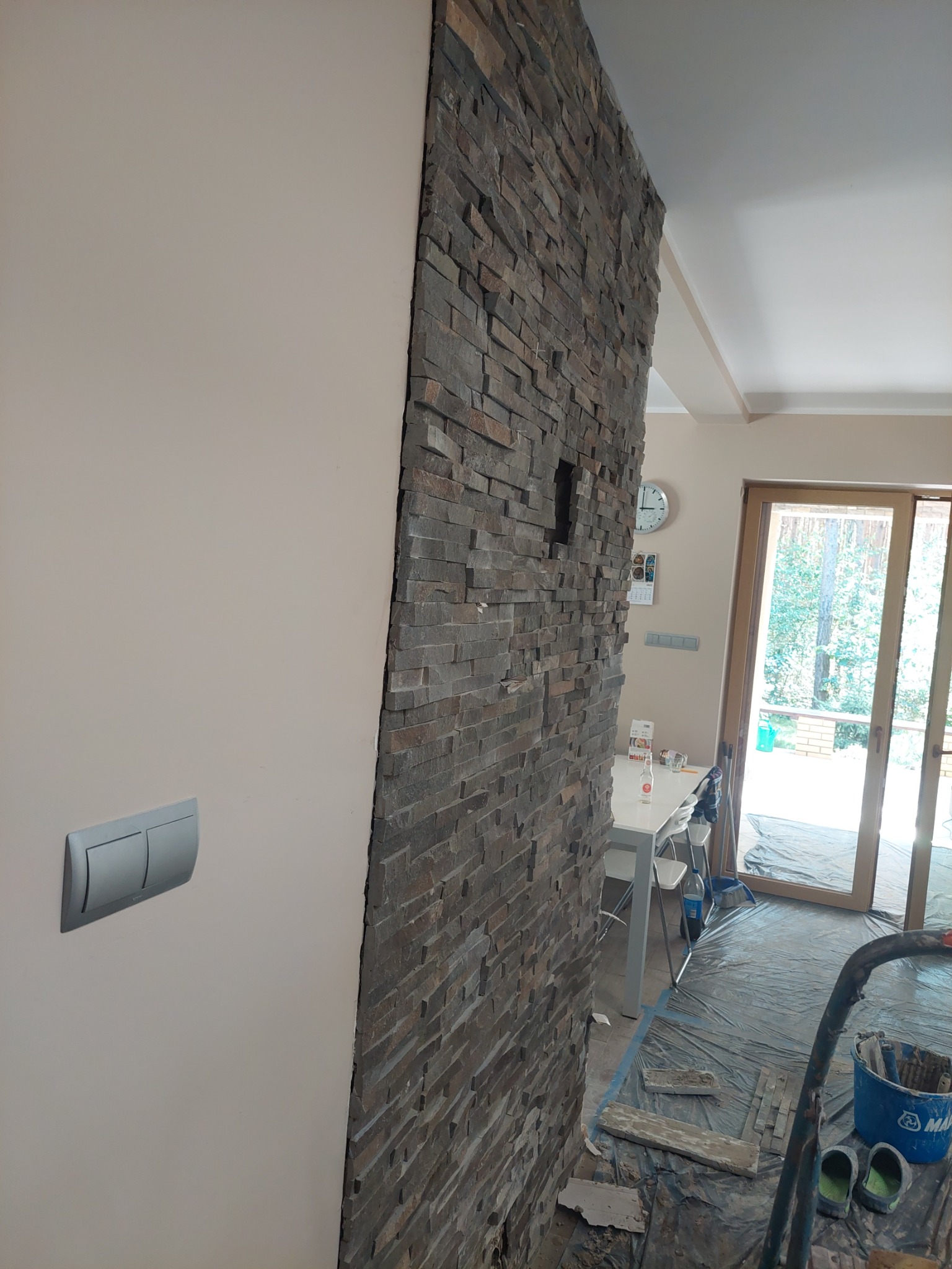 Ściana wyłożona dekoracyjnym kamieniem w odcieniach szarości i brązu, z widocznym fragmentem drugiej ściany pomalowanej na jasny beż, na podłodze folia ochronna i wiadro z narzędziami, sugerujące...