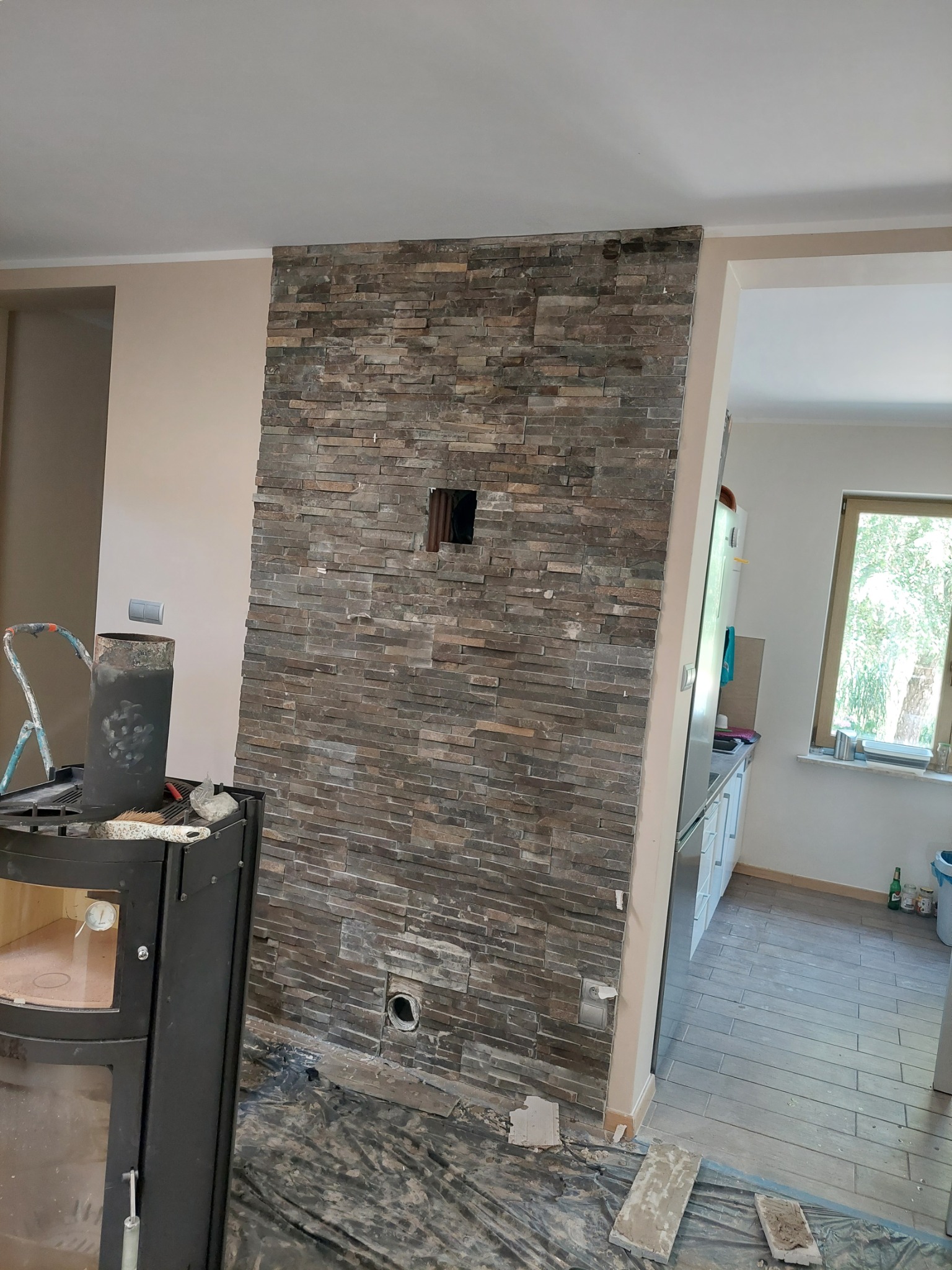 Ściana wyłożona kamieniem dekoracyjnym w trakcie montażu, widoczne otwory na instalacje, fragment kominka i zabezpieczona folią podłoga.