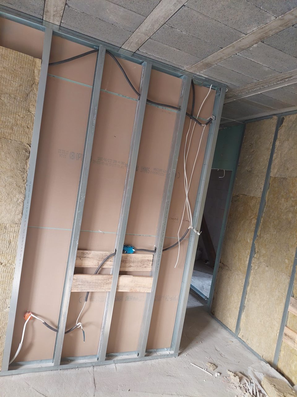 Wnętrze w trakcie remontu: metalowy szkielet ściany działowej z płytami kartonowo-gipsowymi, widoczne przewody elektryczne i izolacja wełną mineralną, surowy betonowy sufit.