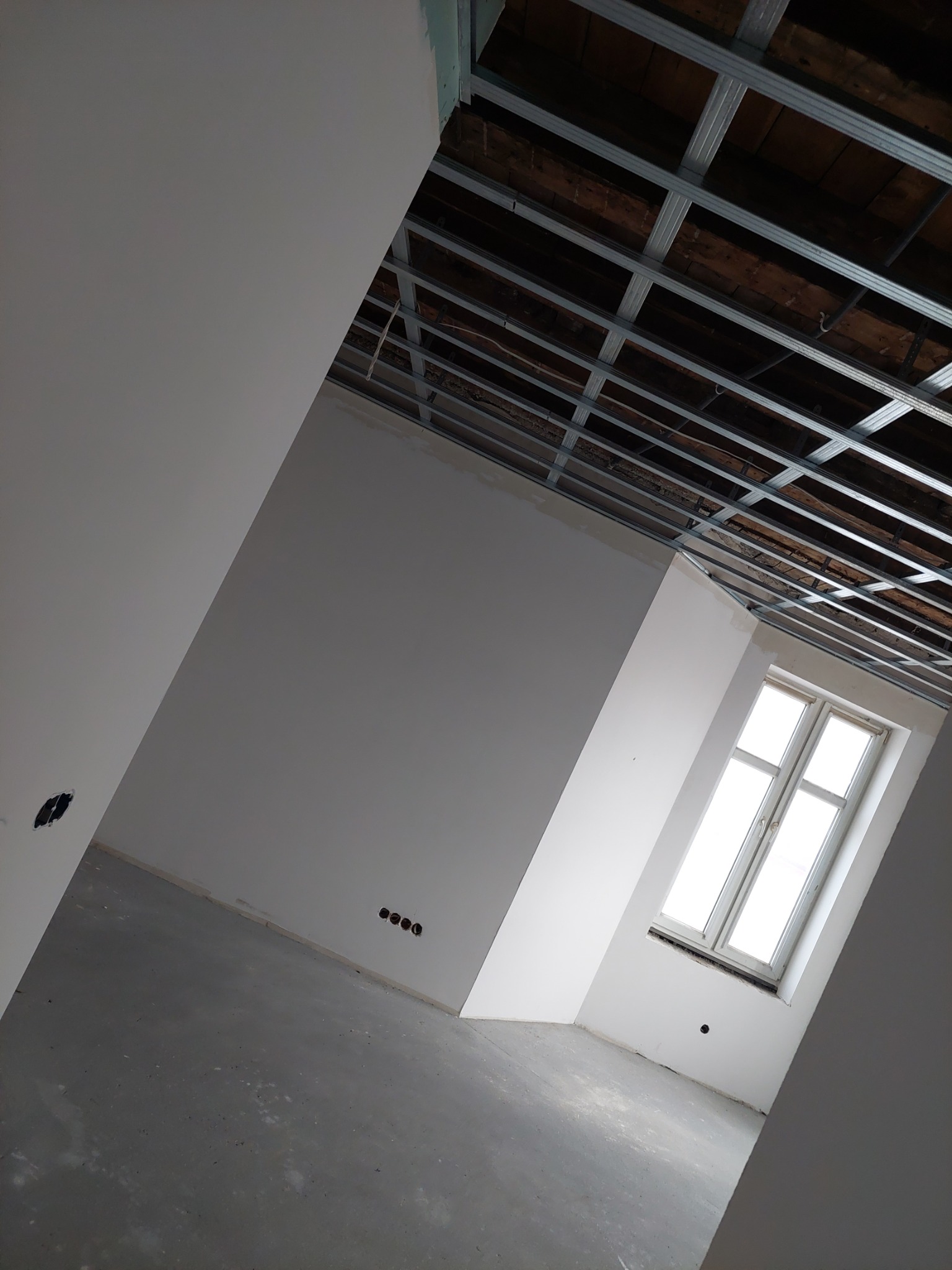 Surowe wnętrze w trakcie prac wykończeniowych: widoczne metalowe profile sufitowe, świeżo położona gładź na ścianach, surowa podłoga i białe okno z widokiem na zewnątrz.