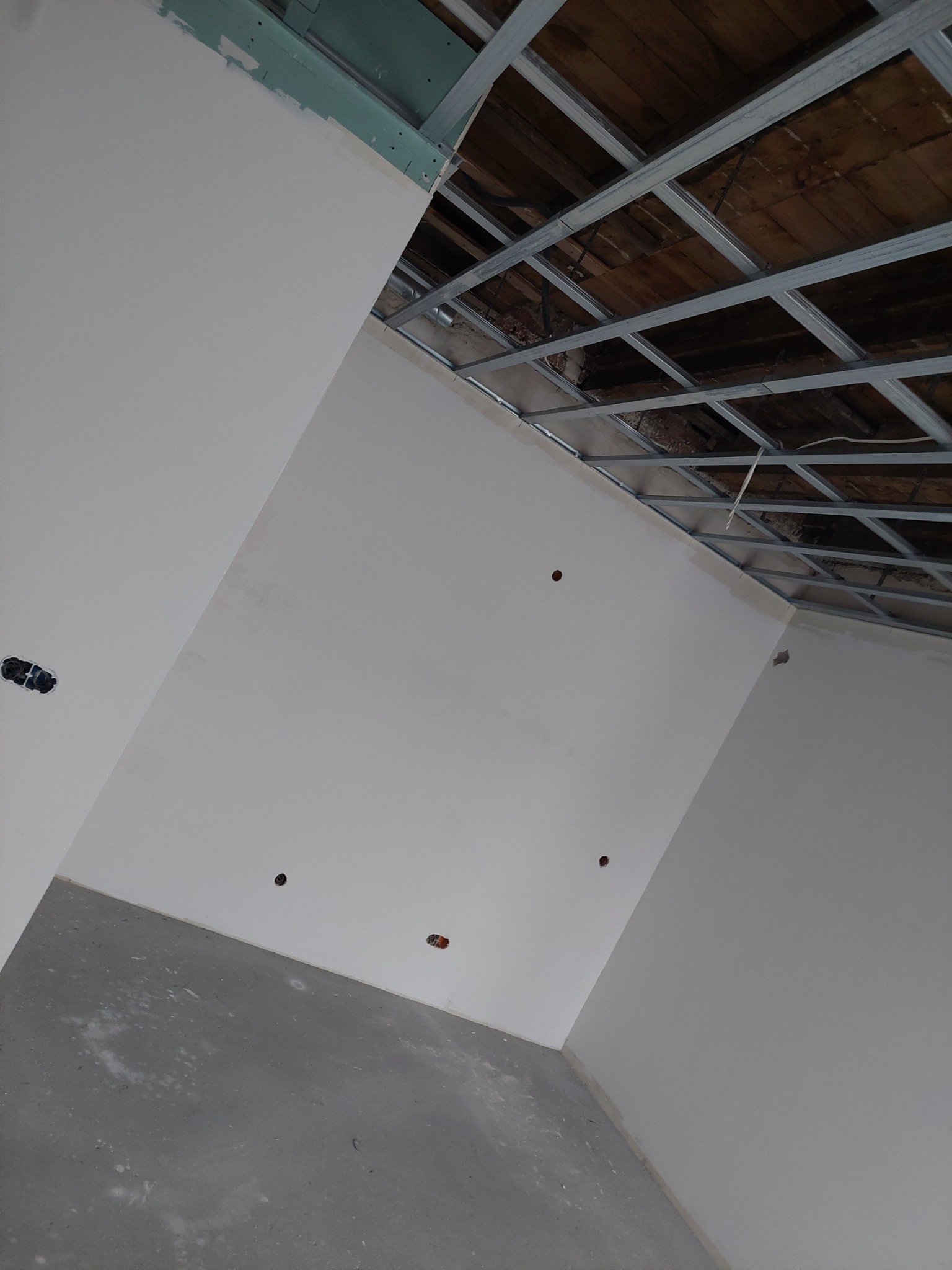 Surowe wnętrze w trakcie remontu: ściany po nałożeniu gładzi, widoczne puszki elektryczne, metalowa konstrukcja sufitu podwieszanego z fragmentami drewnianego stropu.