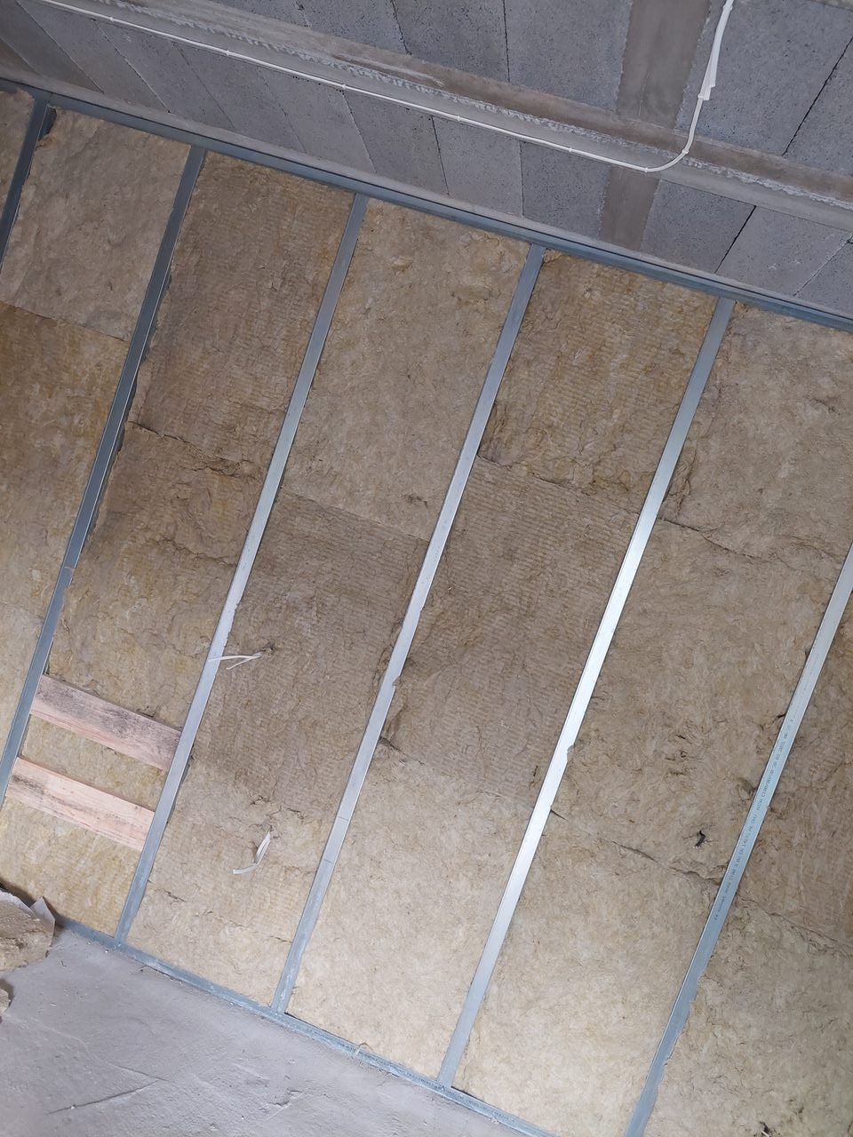 Montaż izolacji z wełny mineralnej w metalowym stelażu na ścianie, widoczne fragmenty surowego sufitu i podłogi.