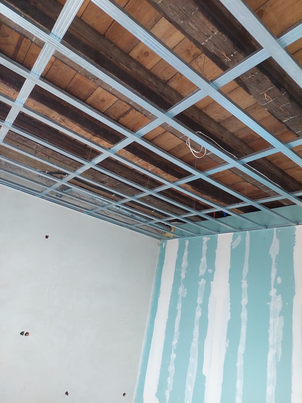 Montaż metalowego rusztu pod sufitem podwieszanym, widoczne stare drewniane belki stropowe i częściowo położona gładź na ścianach.