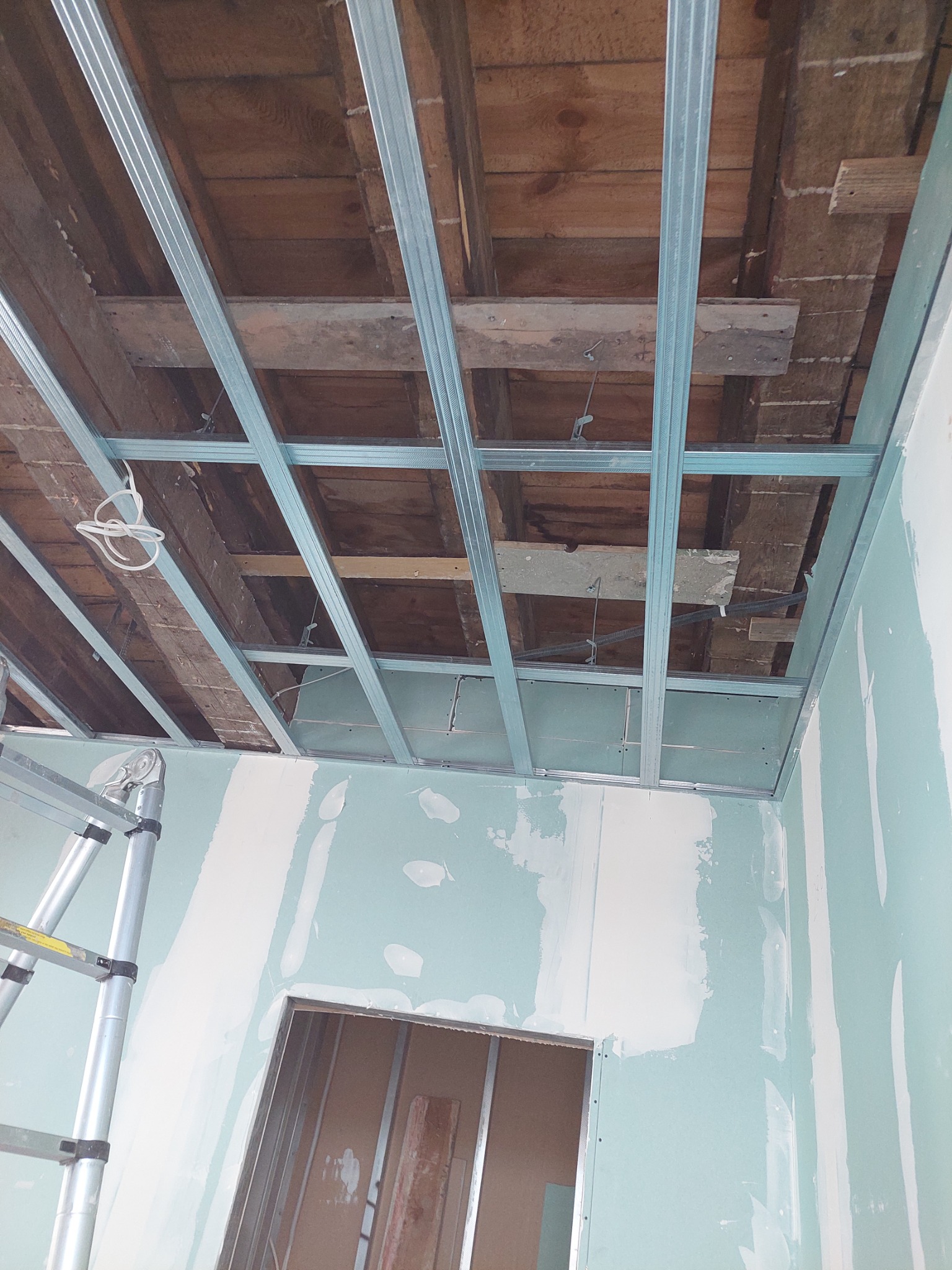 Montaż stelażu sufitu podwieszanego z płyt gipsowo-kartonowych, widoczne drewniane belki stropu i częściowo położone płyty, drabina przystawiona do ściany.