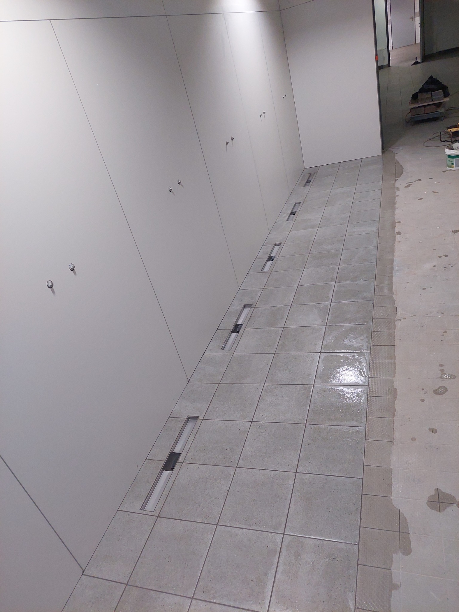 Układanie jasnoszarych płytek podłogowych z wbudowanymi odpływami liniowymi w nowym pomieszczeniu, widoczne ślady zaprawy i przygotowania do dalszych prac wykończeniowych.