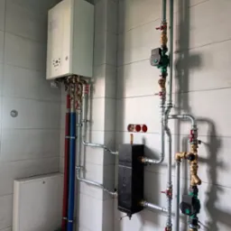 Nowoczesny piec gazowy z rozbudowaną instalacją hydrauliczną, widoczne rury miedziane i stalowe, pompy obiegowe oraz czarny wymiennik ciepła na tle jasnych, ceramicznych płytek ściennych.