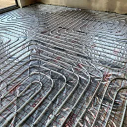 Układanie rur ogrzewania podłogowego na folii refleksyjnej z siatką, widoczne złączki na skraju podłogi i fragment ściany w surowym stanie.