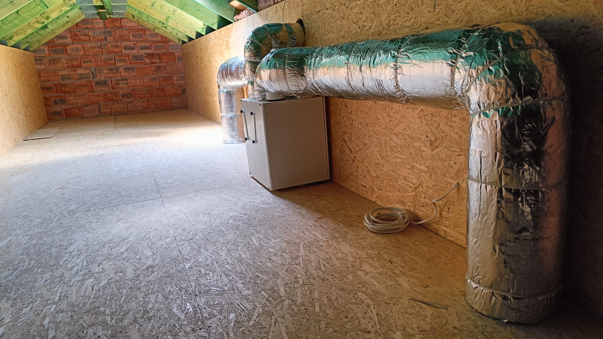 Instalacja rekuperacji w surowym pomieszczeniu na poddaszu, widoczne kanały wentylacyjne owinięte srebrną folią, centrala wentylacyjna, ściany z płyt OSB i fragment ściany z czerwonej cegły.