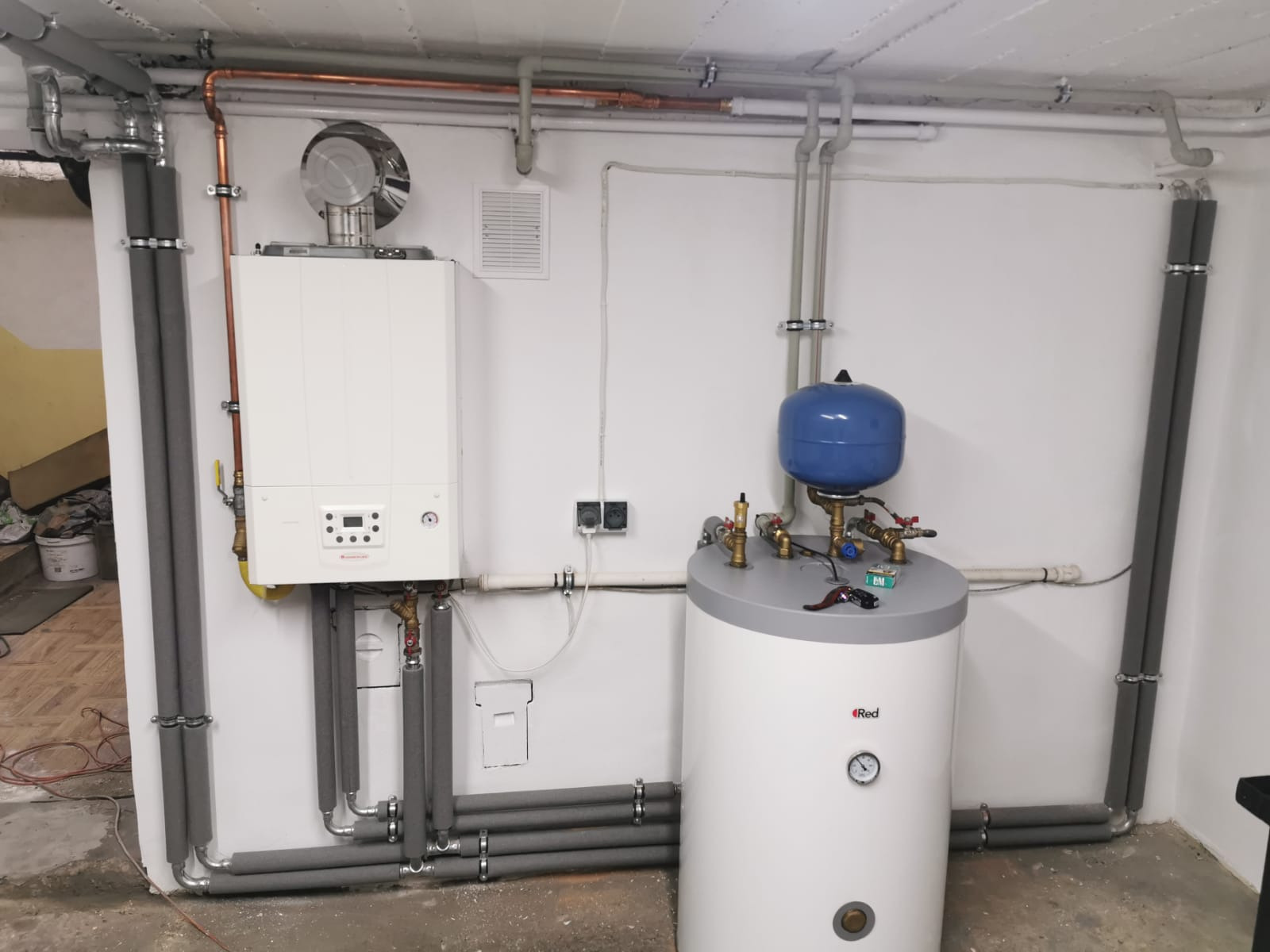 Nowoczesny, biały piec gazowy z podłączonym zasobnikiem ciepłej wody z niebieskim zbiornikiem wyrównawczym, widoczne rury w izolacji termicznej, na ścianie kratka wentylacyjna i gniazdka elektryczne.