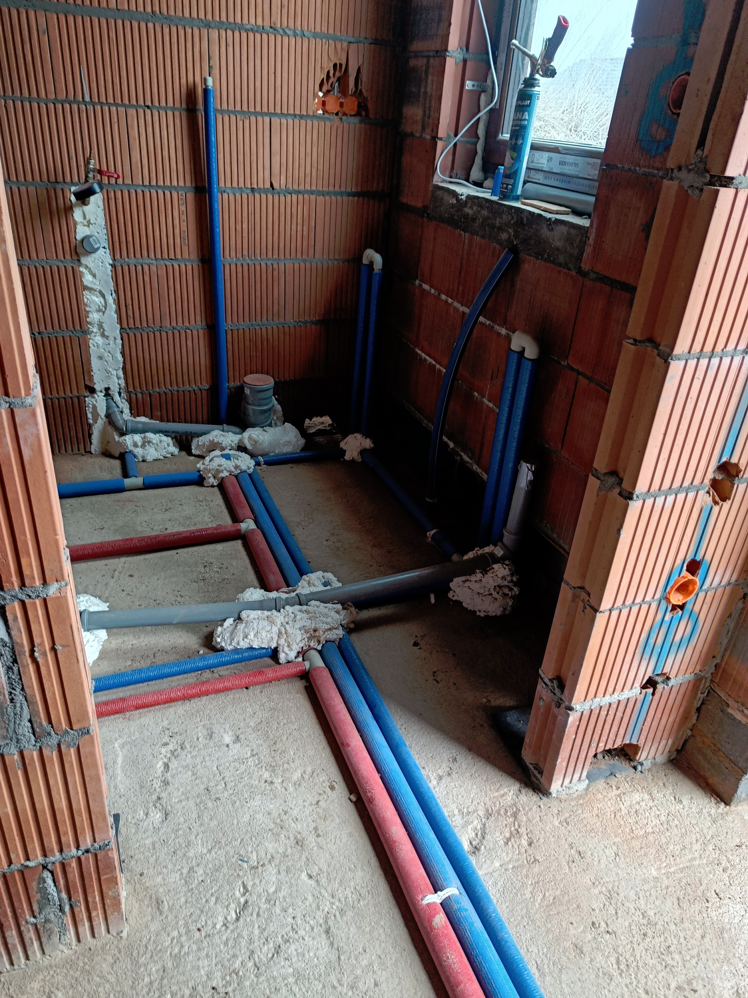 Wnętrze w trakcie budowy z widocznymi instalacjami wodnymi i grzewczymi, rury w kolorach niebieskim i czerwonym, zabezpieczone pianką montażową, ściany z czerwonej cegły kratówki.