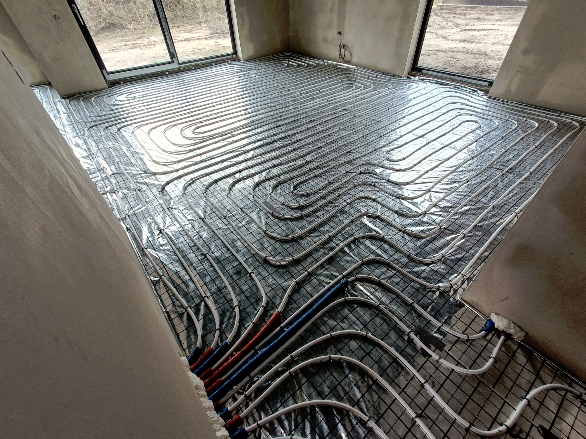 Rozłożone rury ogrzewania podłogowego na folii izolacyjnej z widocznymi czerwonymi i niebieskimi rurami zasilającymi, w surowym wnętrzu budynku z oknami.