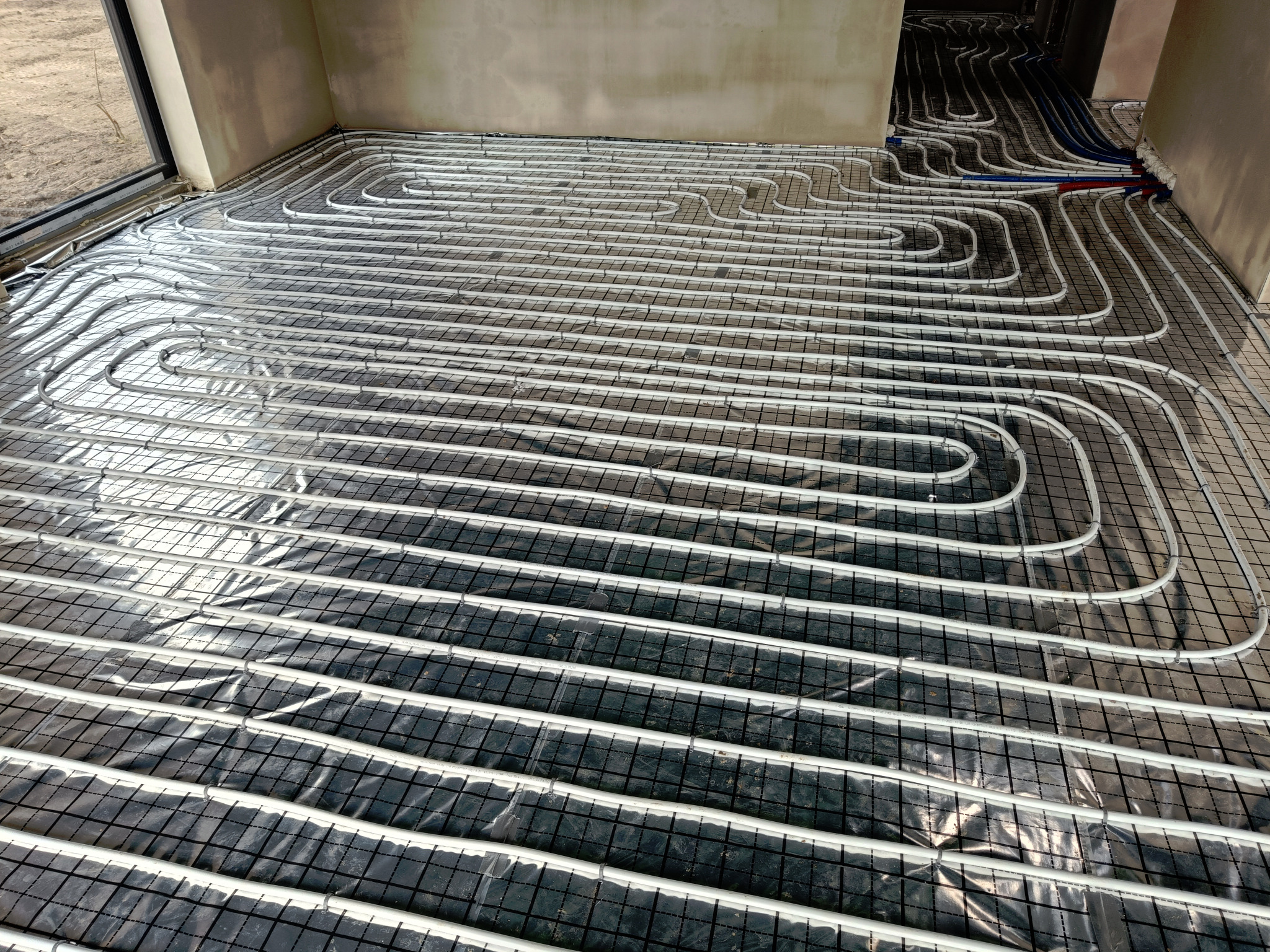 Rozłożona instalacja ogrzewania podłogowego z białych rurek na czarnej siatce, widoczne podłączenia rur w oddali, surowy stan wykończenia wnętrza.