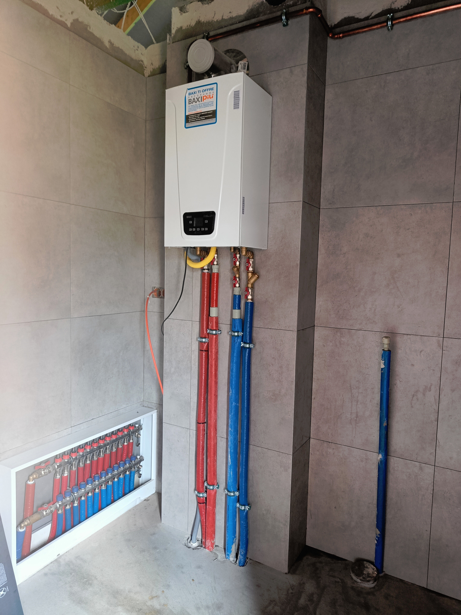 Nowy, biały piec gazowy Baxi, zamontowany na szarej, kafelkowanej ścianie, z widocznymi rurami w izolacji czerwonej i niebieskiej, oraz rozdzielacz ogrzewania podłogowego w białej szafce.