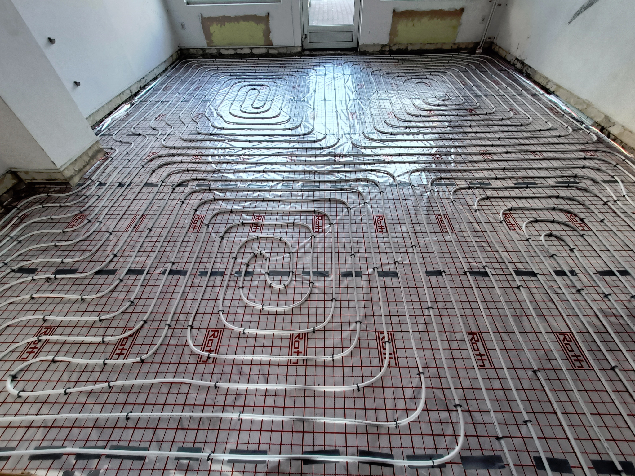 Białe rurki ogrzewania podłogowego ułożone na podkładzie z czerwoną kratką, widoczne mocowania rurek, surowy stan wykończenia pomieszczenia w tle.