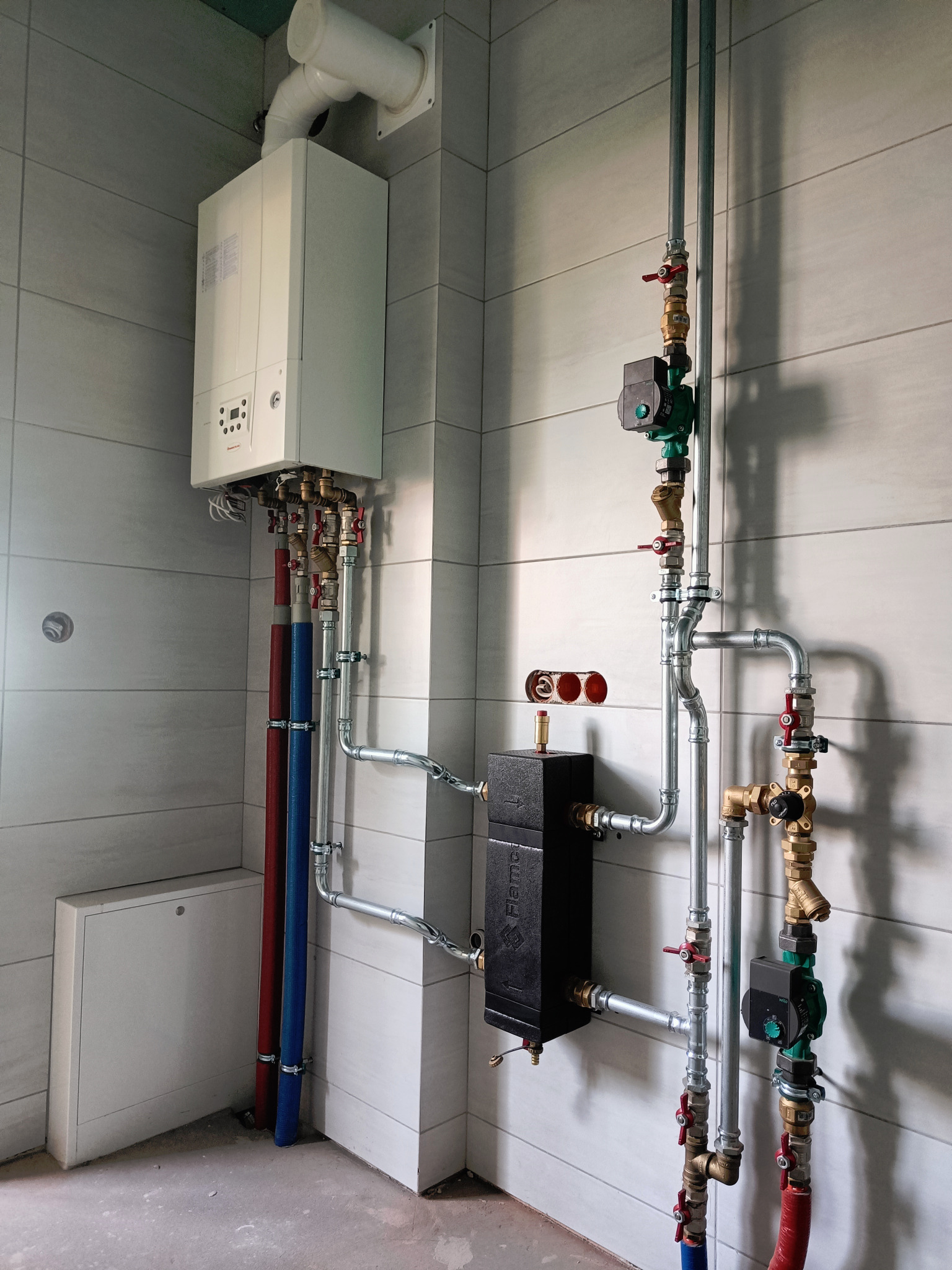 Nowoczesny piec gazowy z rozbudowaną instalacją hydrauliczną, widoczne rury miedziane i stalowe, pompy obiegowe oraz czarny wymiennik ciepła na tle jasnych, ceramicznych płytek ściennych.