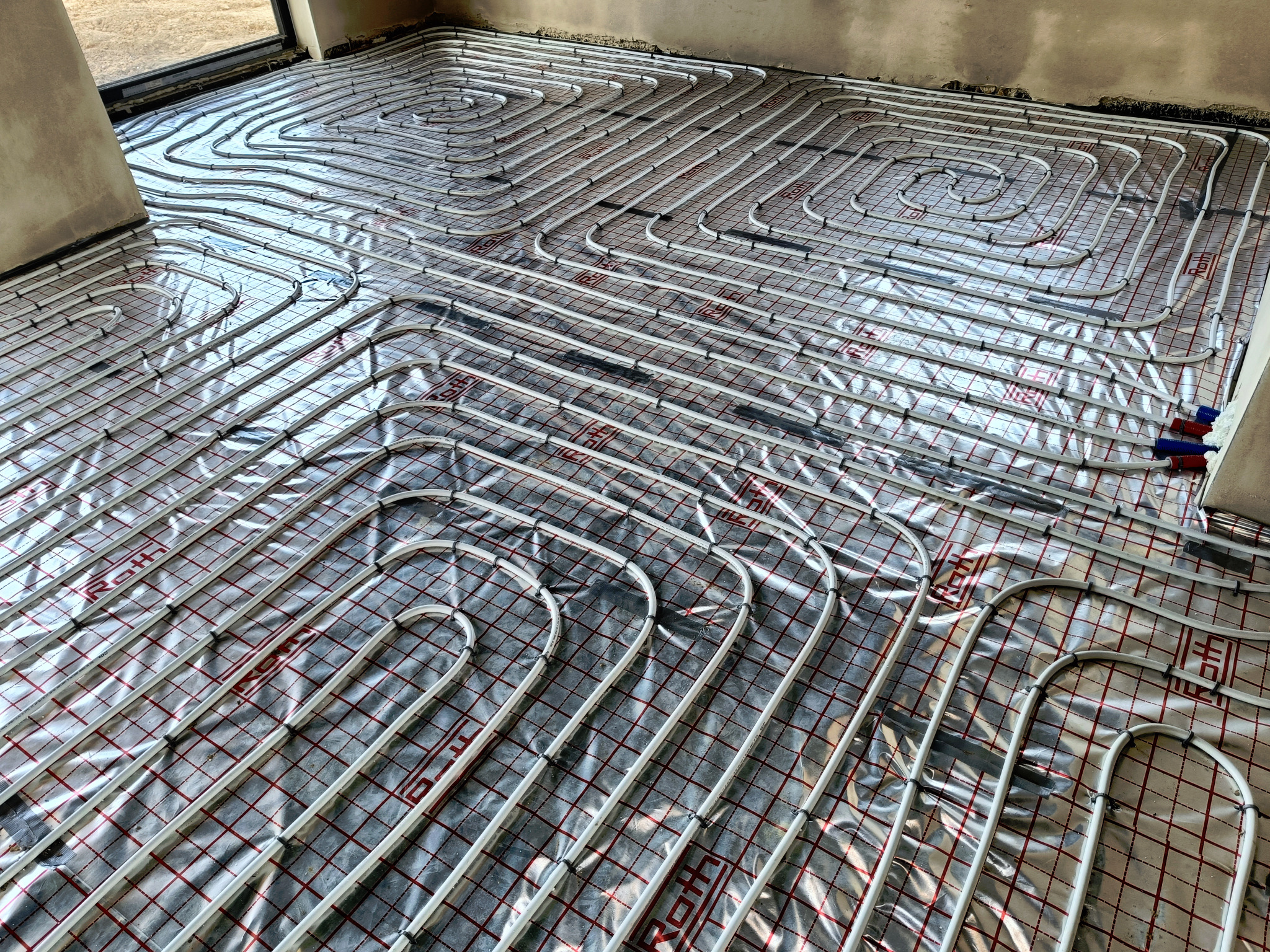 Układanie rur ogrzewania podłogowego na folii refleksyjnej z siatką, widoczne złączki na skraju podłogi i fragment ściany w surowym stanie.