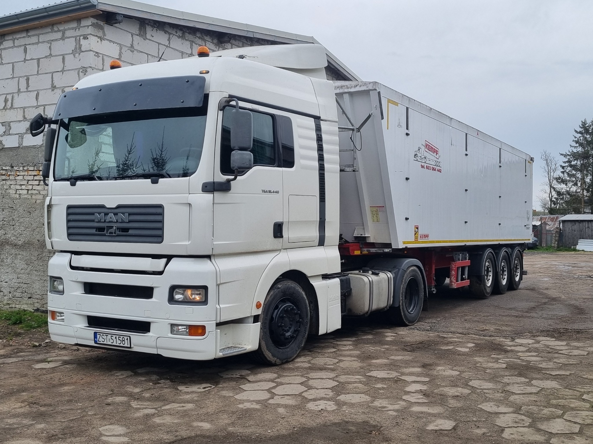 Biały ciągnik siodłowy MAN z naczepą wywrotką marki Kempf, zaparkowany na placu z betonową kostką, widoczne numery rejestracyjne ZST 51581 i logo producenta naczepy.