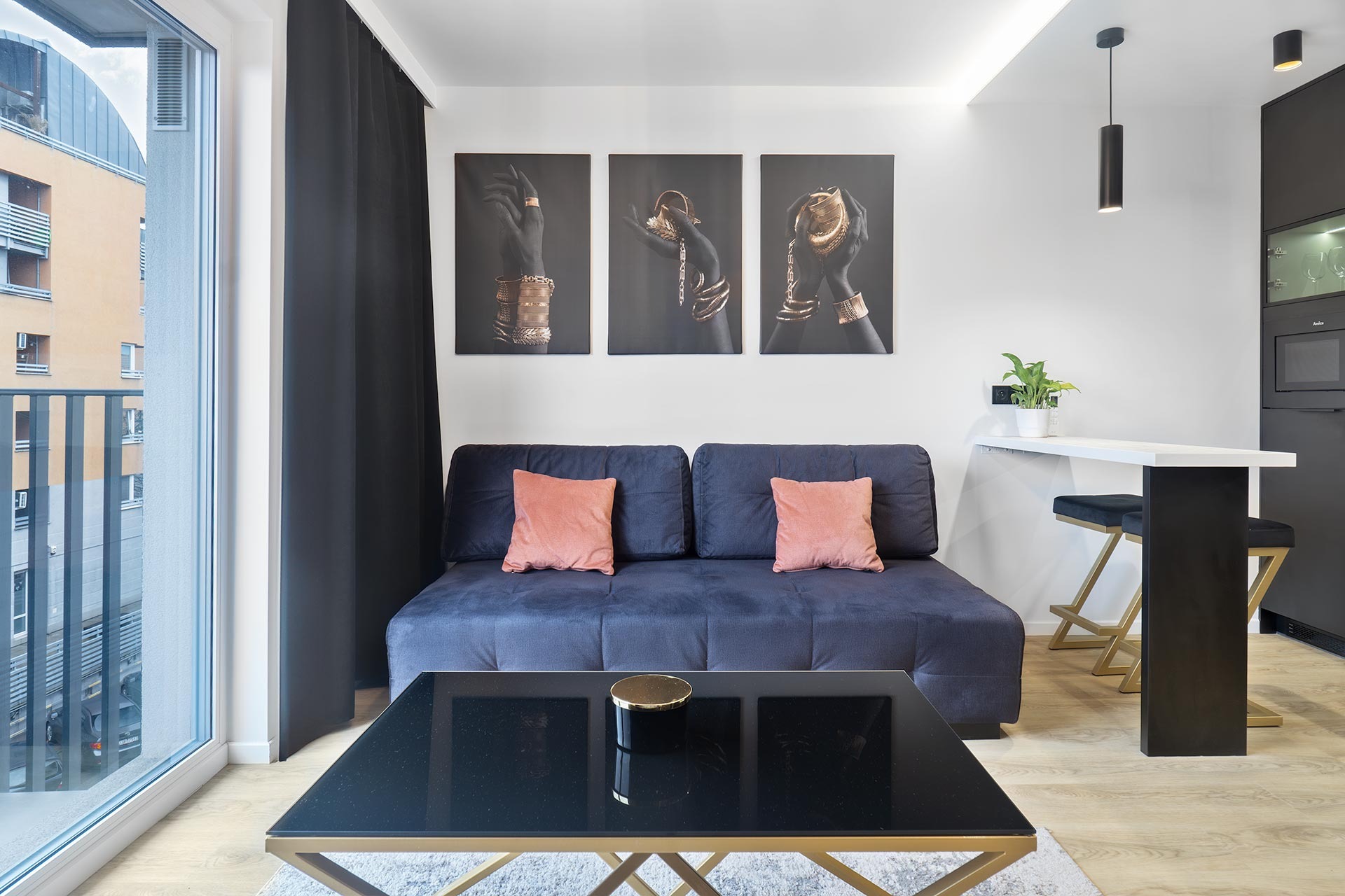 Nowoczesny salon z ciemnoniebieską sofą z brzoskwiniowymi poduszkami, czarnym stolikiem kawowym ze złotymi nogami, tryptykiem przedstawiającym dłonie w złotej biżuterii, czarnymi zasłonami...
