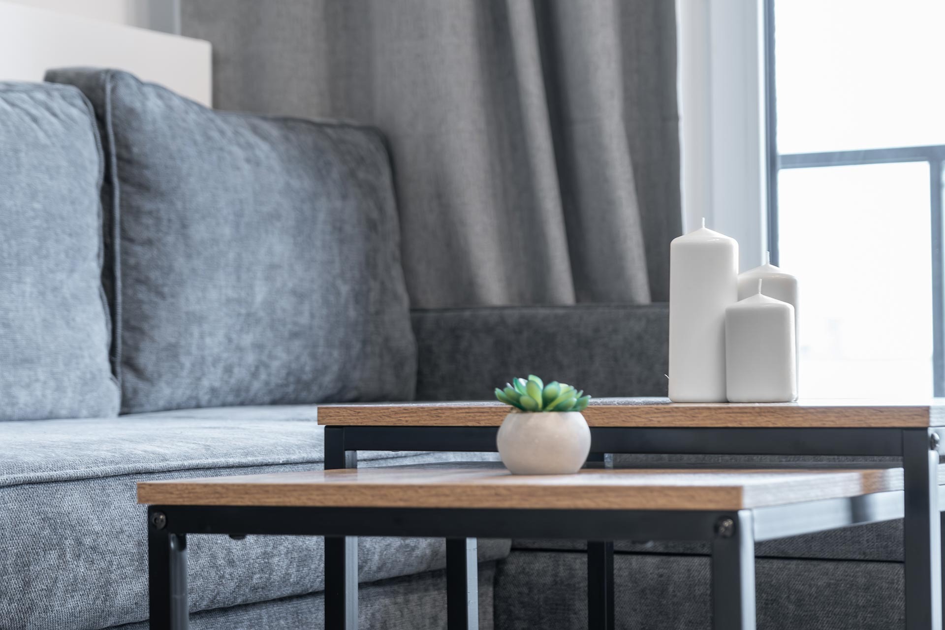 Jasnoszara sofa z poduszkami, stolik kawowy z drewnianym blatem i czarną metalową ramą, trzy białe świece i mały sukulent w beżowej doniczce, wszystko w jasnym, nowoczesnym wnętrzu.