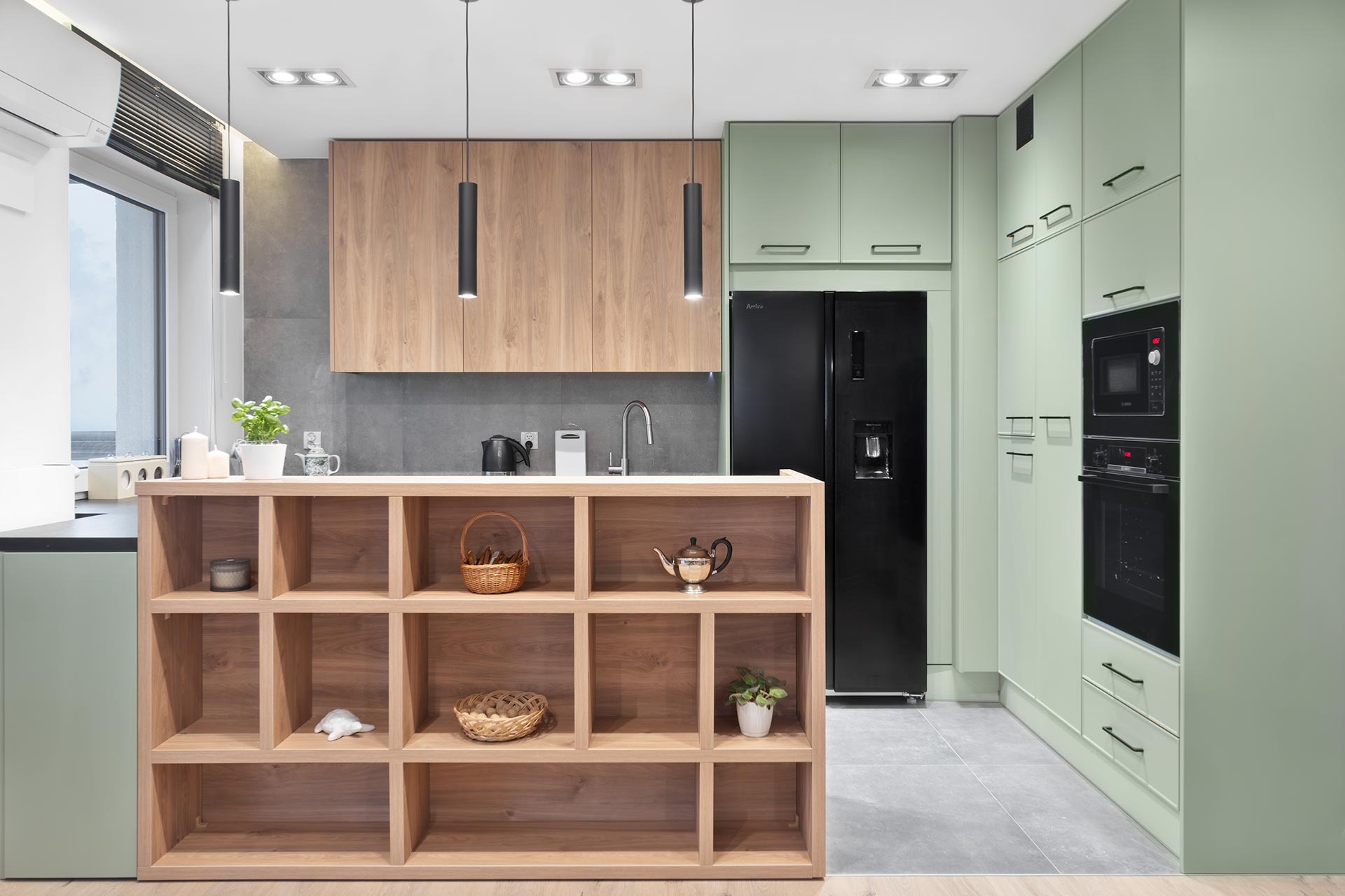 Nowoczesna kuchnia z drewnianą wyspą, miętowe szafki i czarny sprzęt AGD, oświetlenie punktowe i wiszące lampy cylindryczne.