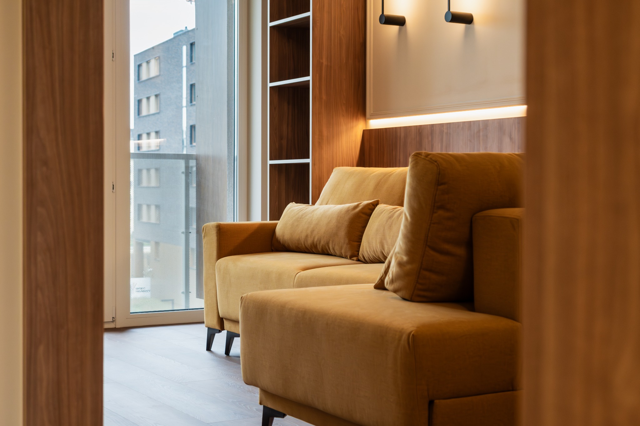 Oświetlony fragment salonu z aksamitną, musztardową sofą i widokiem na budynek mieszkalny za oknem.