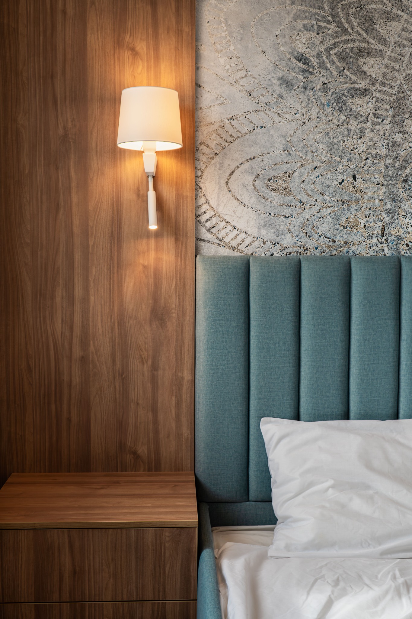 Nowoczesna sypialnia: drewniana szafka nocna z lampką, turkusowe wezgłowie z pionowymi przeszyciami oraz dekoracyjna szara tapeta z abstrakcyjnym wzorem.