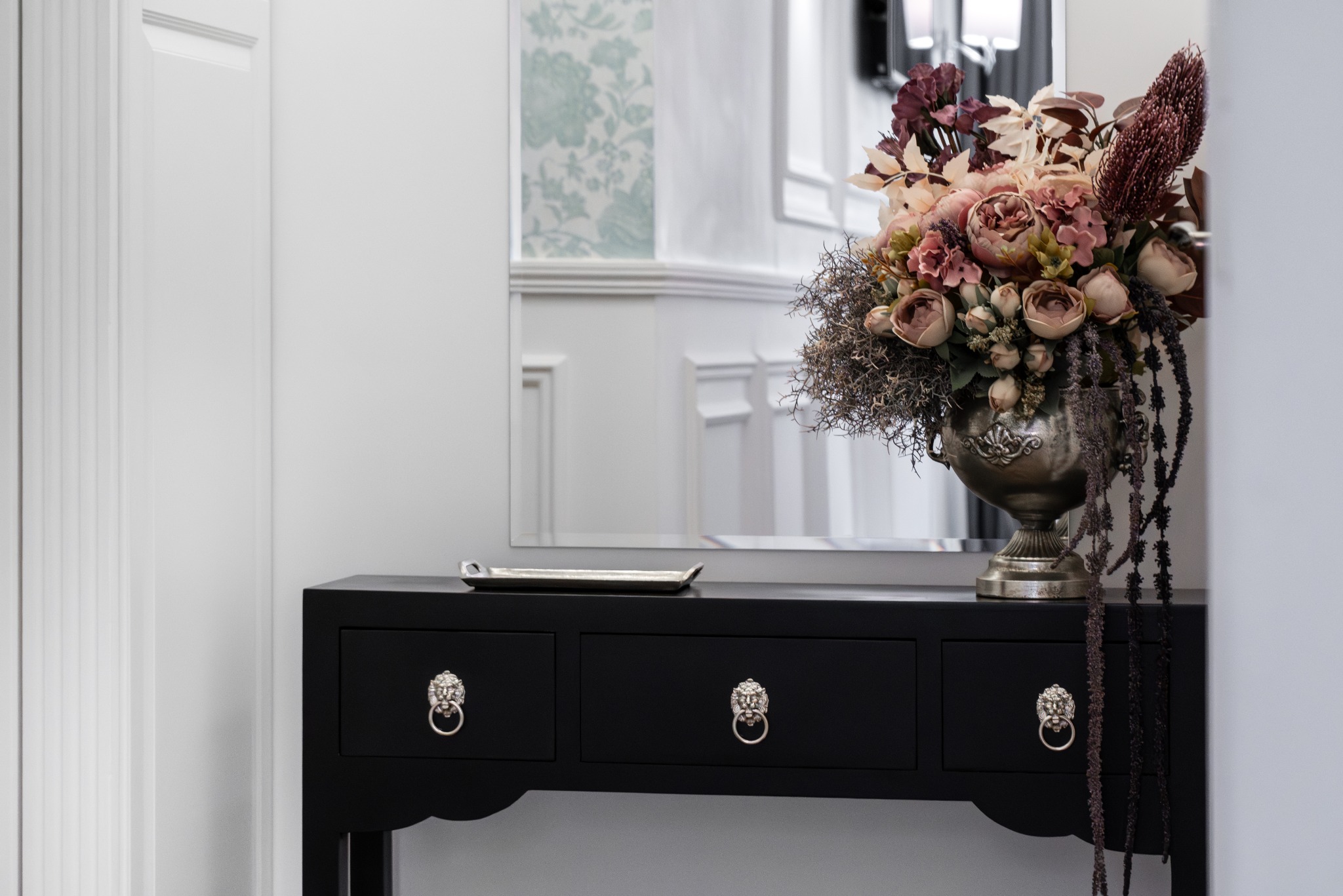 Czarna konsola z trzema szufladami i uchwytami w kształcie lwich głów, srebrna taca, lustro i kompozycja kwiatowa z różowych i bordowych kwiatów w metalowym wazonie.