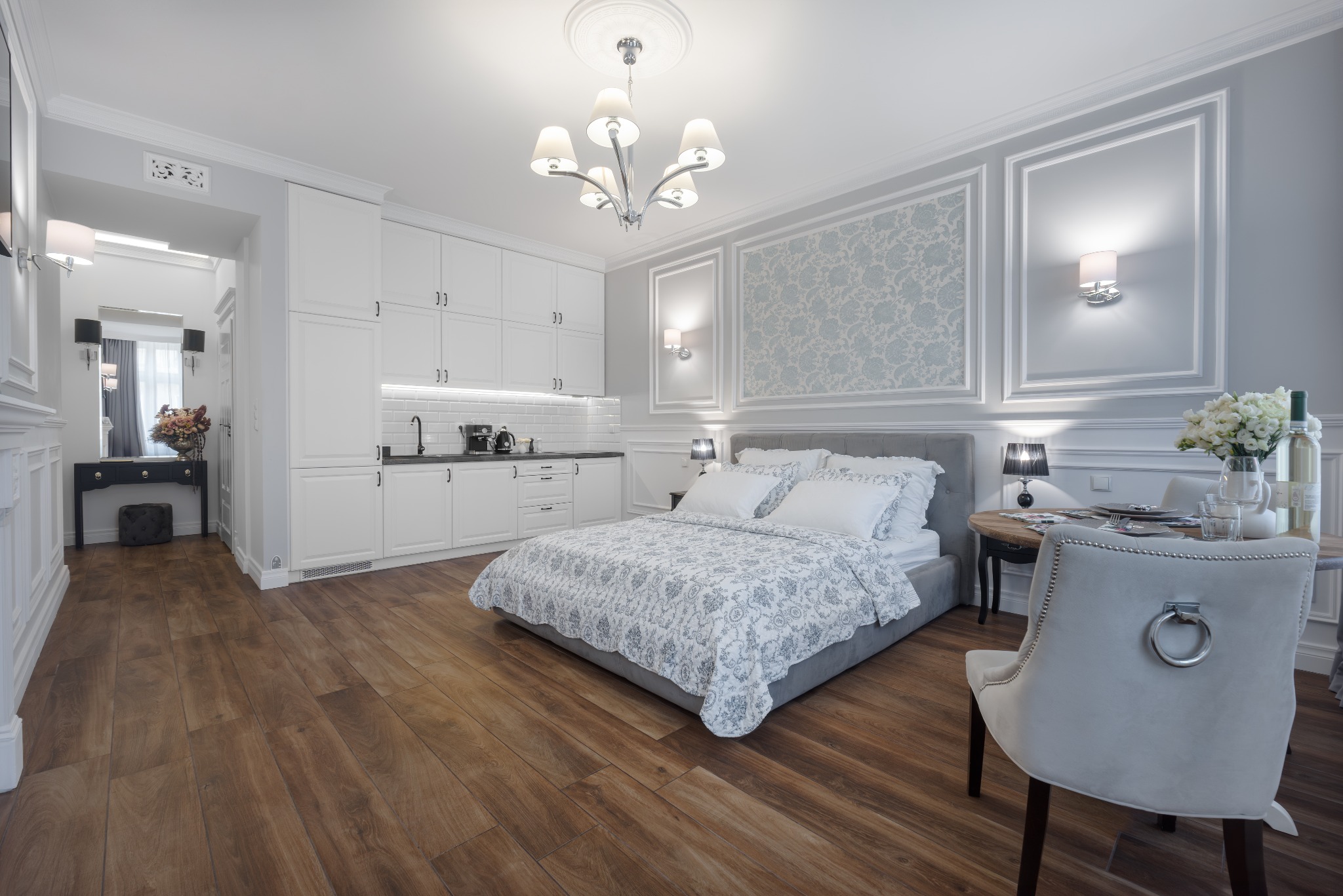 Eleganckie studio z sypialnią i aneksem kuchennym w stylu klasycznym: drewniana podłoga, szara tapicerowana rama łóżka, białe meble kuchenne, okrągły stół z nakryciem i bukietem kwiatów.
