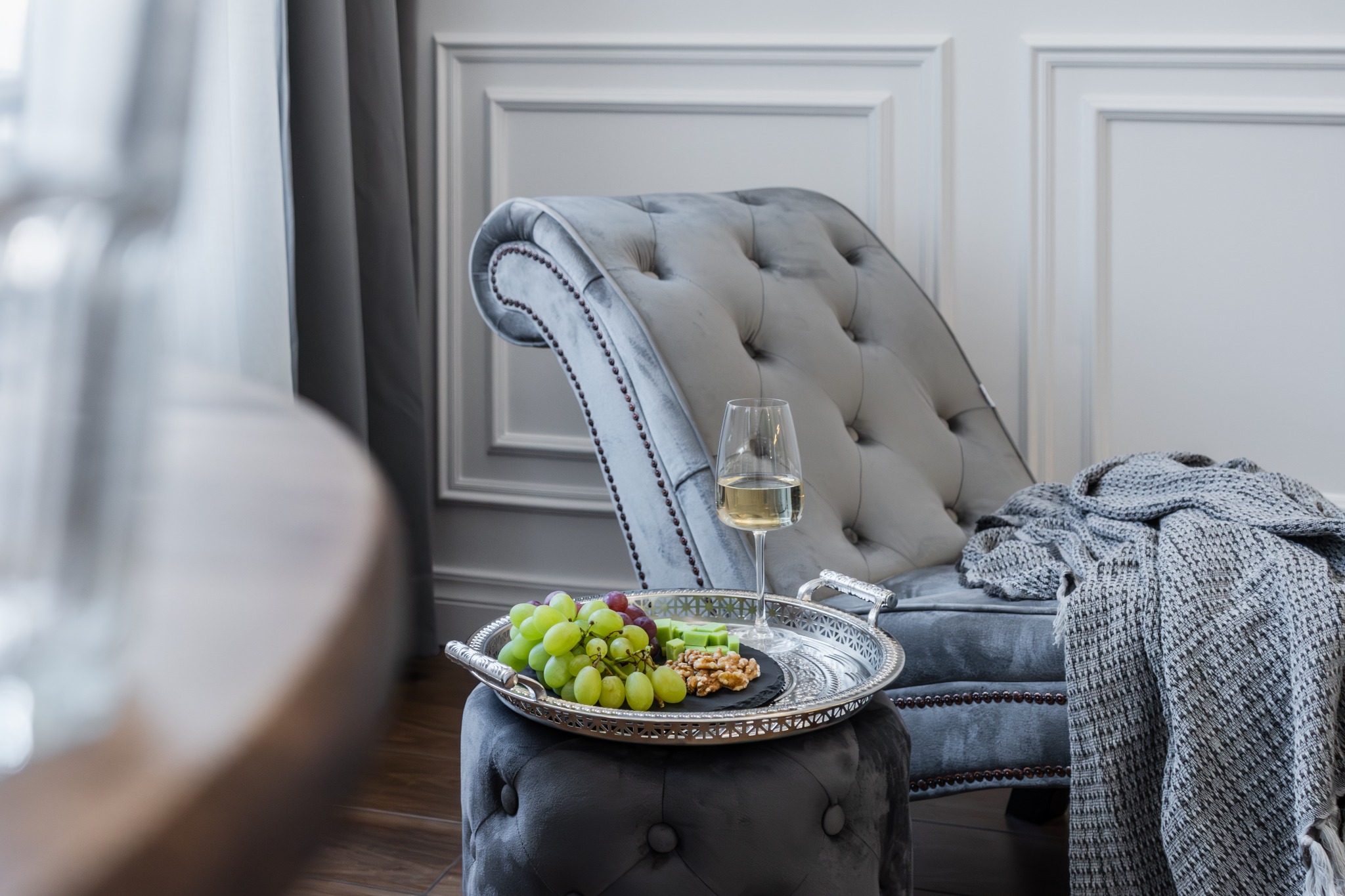 Elegancki szary fotel z aksamitnym obiciem, ozdobiony ćwiekami, przykryty pledem, obok srebrna taca z winogronami, serem i orzechami, kieliszek białego wina, w tle sztukateria na ścianie i zasłony.