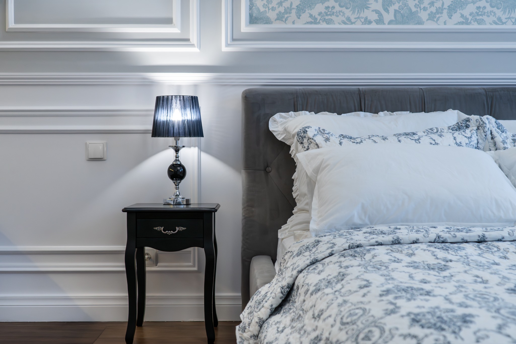 Elegancka sypialnia z szarym zagłówkiem, wzorzystą pościelą i czarną szafką nocną z lampką, oświetlona delikatnym światłem.