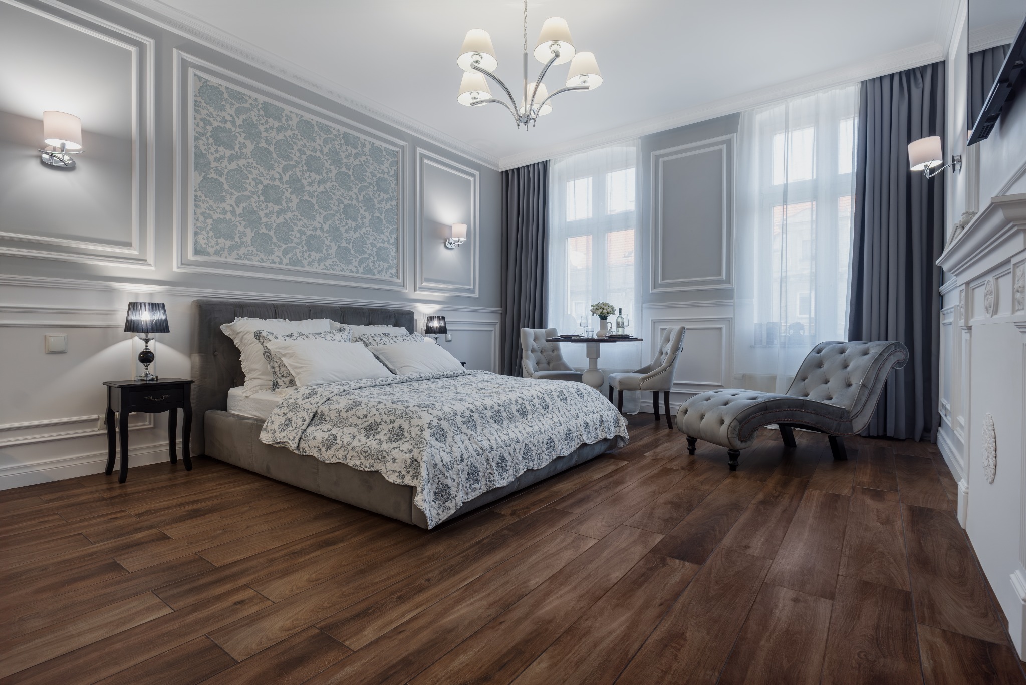 Elegancka sypialnia z łóżkiem typu king-size, drewnianą podłogą, sztukaterią na ścianach, żyrandolem i małym stolikiem jadalnym z krzesłami w tle.