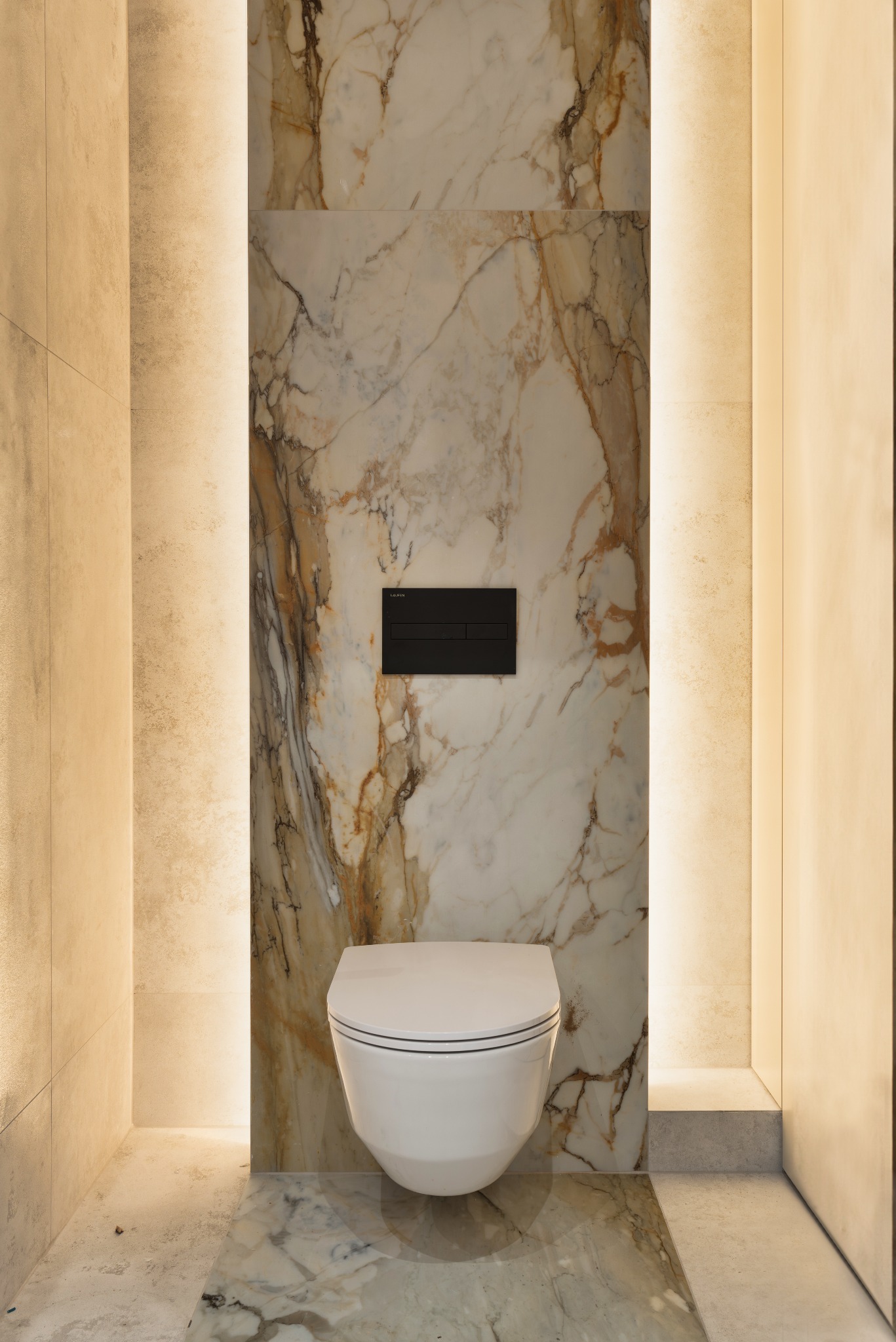 Nowoczesna toaleta podwieszana z czarnym przyciskiem spłukującym na ścianie z marmurowym wzorem, oświetlona z obu stron ciepłym światłem.