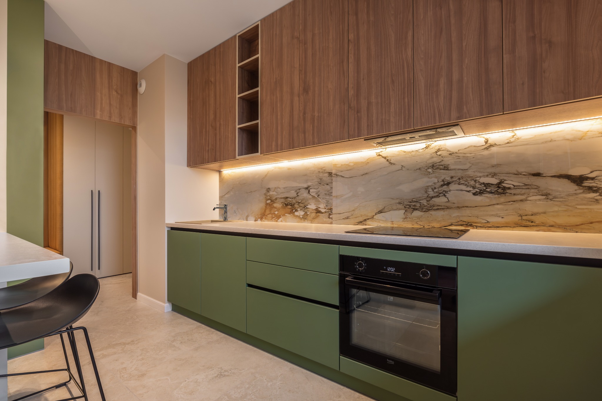 Nowoczesna kuchnia z zielonymi szafkami, marmurowym backsplashiem podświetlonym LED, drewnianymi szafkami górnymi i czarnymi hokerami przy białym blacie.