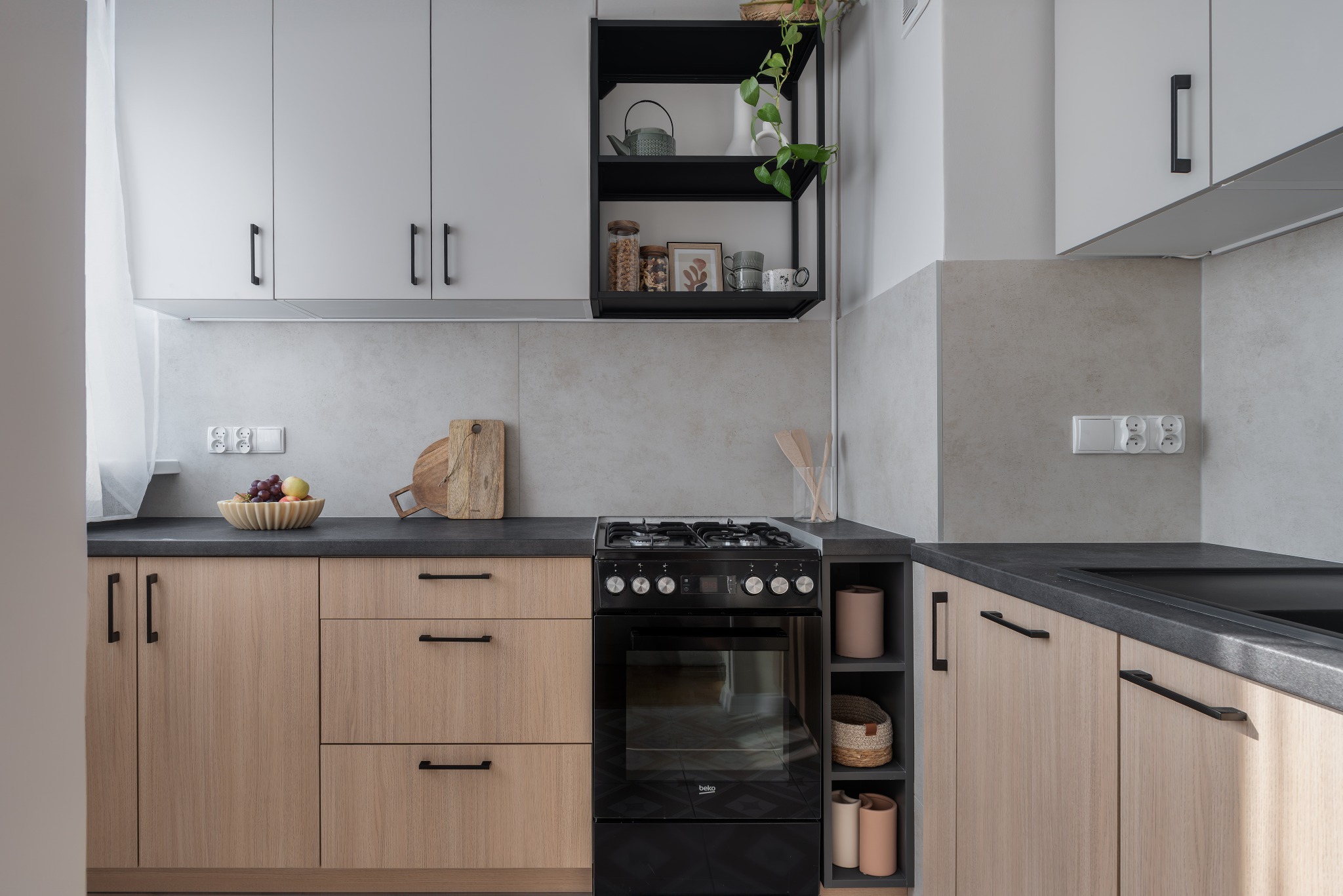 Jasna kuchnia z drewnianymi frontami szafek, czarnym blatem i piekarnikiem, uzupełniona o czarną półkę z dekoracjami i rośliną.
