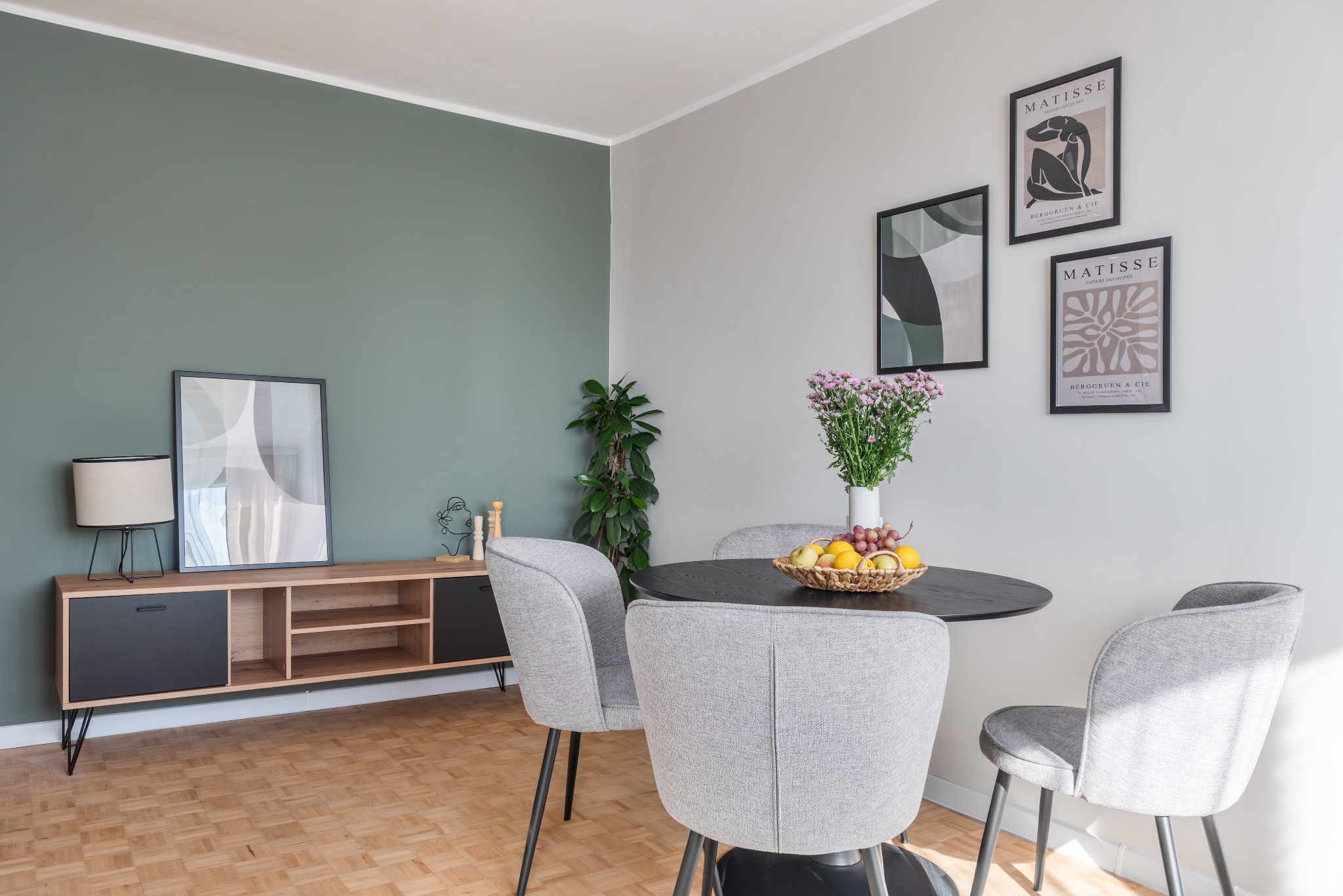 Jasne wnętrze jadalni z okrągłym, czarnym stołem, szarymi krzesłami, bukietem kwiatów w wazonie i koszem owoców. Na ścianie wiszą trzy grafiki w czarnych ramkach, a obok znajduje się drewniana...