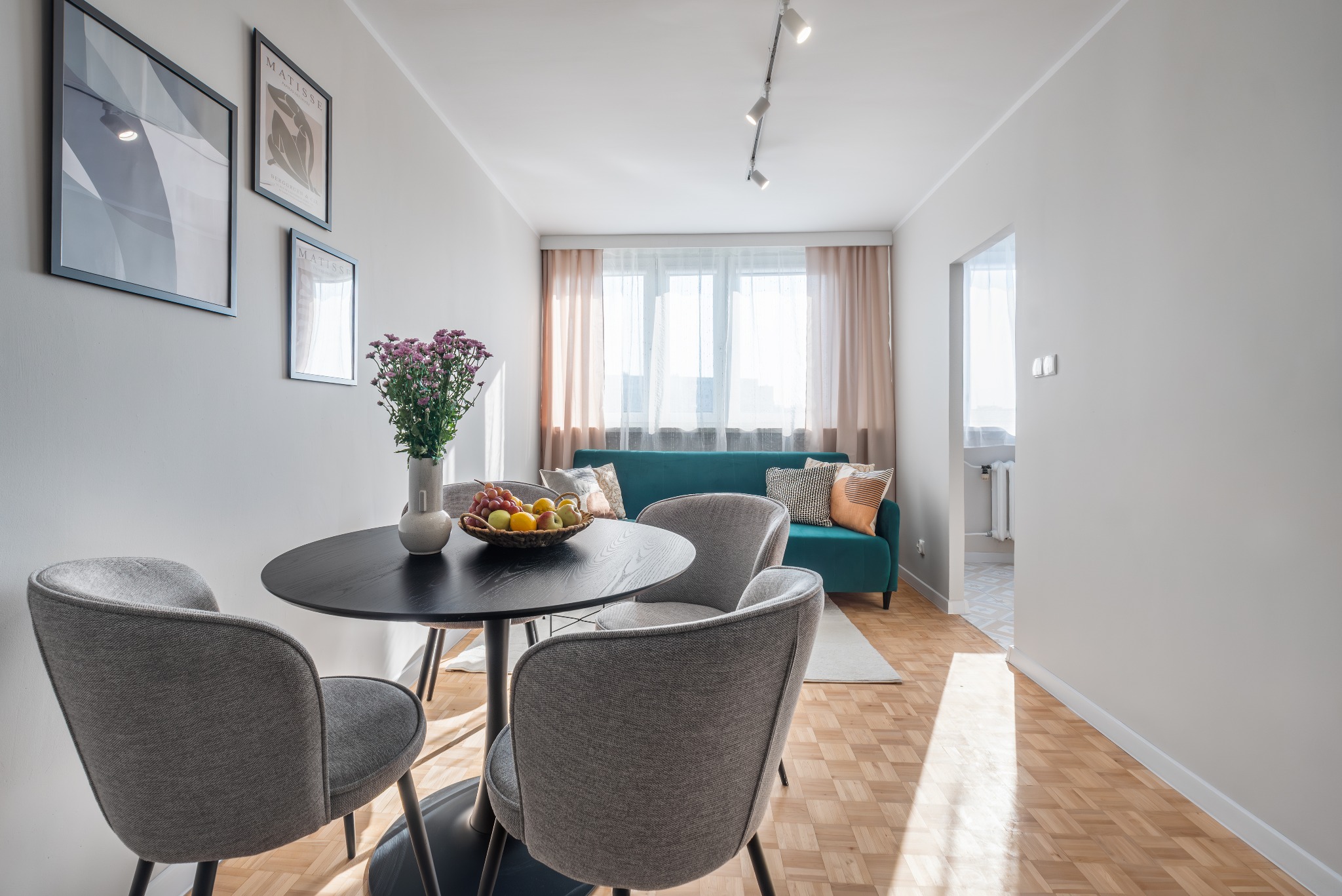 Jasne wnętrze mieszkania z okrągłym, czarnym stołem, szarymi krzesłami, bukietem kwiatów w wazonie i turkusową sofą w tle, oświetlone przez reflektory na suficie i światło dzienne wpadające przez...