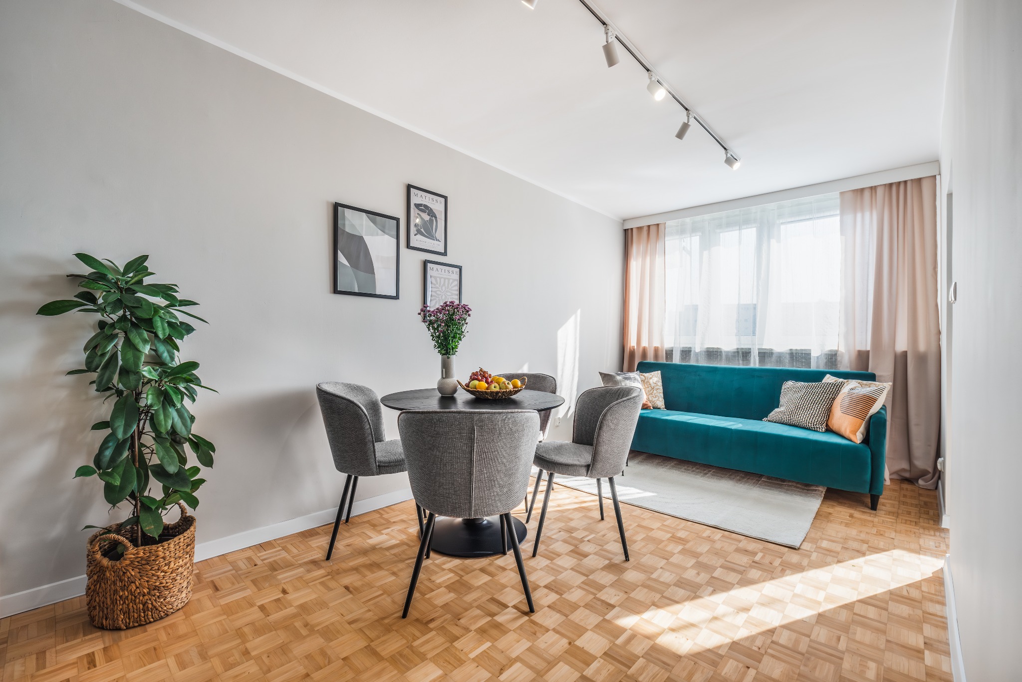 Jasne wnętrze z okrągłym stołem, szarymi krzesłami, turkusową sofą z poduszkami, drewnianą podłogą parkietową i rośliną doniczkową w wiklinowym koszu.