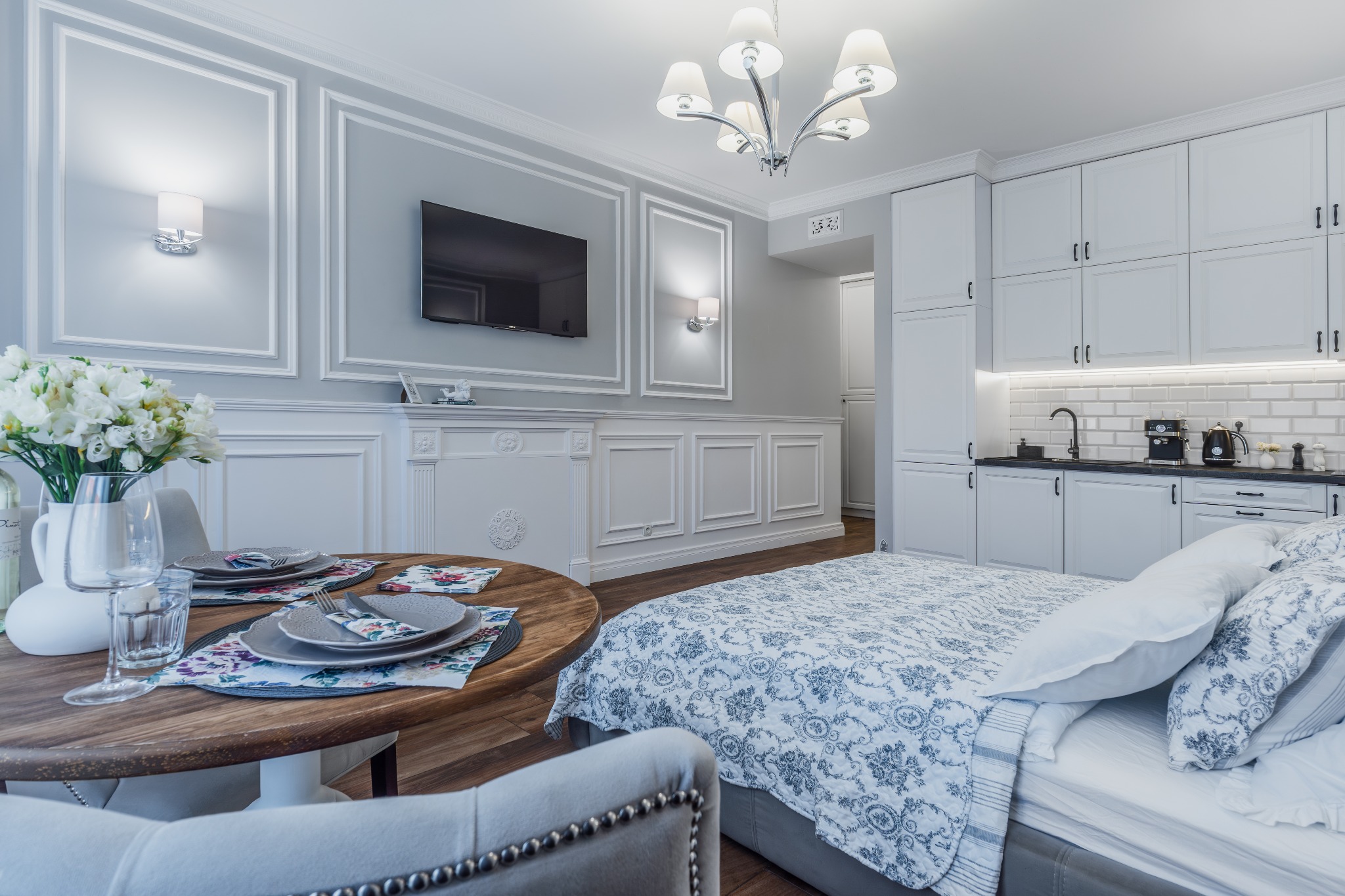 Nowoczesne studio z aneksem kuchennym, okrągłym stołem z nakryciem i łóżkiem z kwiatowym wzorem pościeli, ściany w białej sztukaterii, telewizor i lampy ścienne.