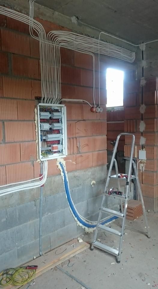 Rozdzielnica elektryczna z licznymi przewodami w białych peszlach, zamontowana na ścianie z czerwonej cegły, z niebieskim peszlem doprowadzającym kable od dołu, pianka montażowa uszczelniająca...