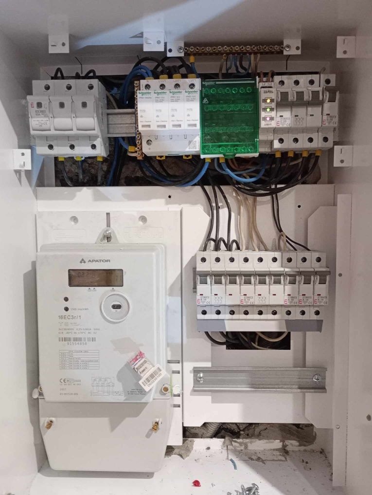 Wnętrze skrzynki elektrycznej z licznikiem, bezpiecznikami i przewodami, widoczne elementy instalacji elektrycznej.