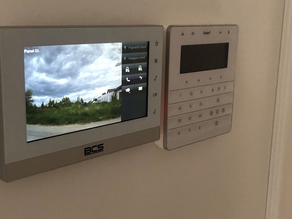Zamontowany na ścianie panel wideodomofonu marki BCS z widocznym podglądem z kamery na posesję z budynkiem w tle, obok klawiatura numeryczna domofonu marki Salef.