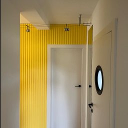 Michał Kromka - Wnętrze korytarza z żółtą, teksturowaną ścianą i białymi drzwiami z okrągłym okienkiem. Widoczne elementy oświetlenia na suficie i drewniana podłoga.