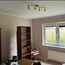 Michał Kromka - Beżowe wnętrze pokoju z brązowym regałem, biurkiem, krzesłem i białym grzejnikiem pod oknem. Ściany świeżo pomalowane na jednolity kolor.