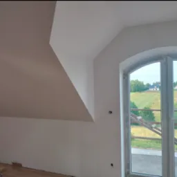 Wnętrze po malowaniu: białe ściany poddasza ze skosami i oknem łukowym z widokiem na zielone wzgórza. Częściowo widoczna podłoga w trakcie montażu.